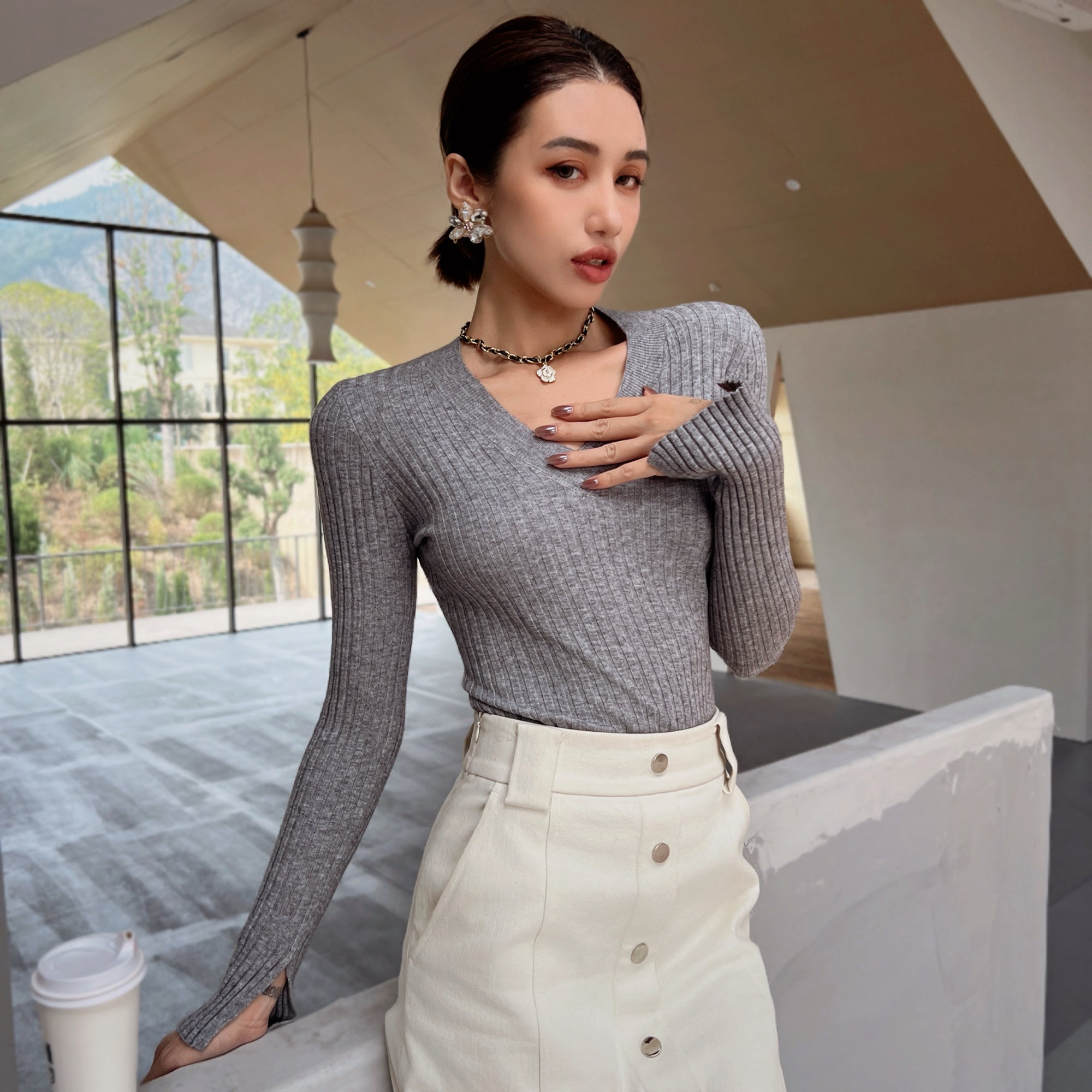 A woman wearing a gray sweater and white skirt stands in a room with large windows, holding a cup of coffee.