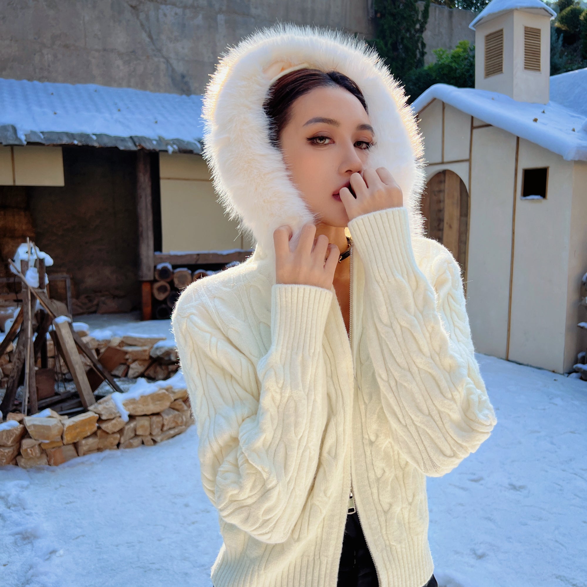 A woman wearing a white fur-lined hooded sweater stands in front of a snowy building, with her hands covering her mouth.