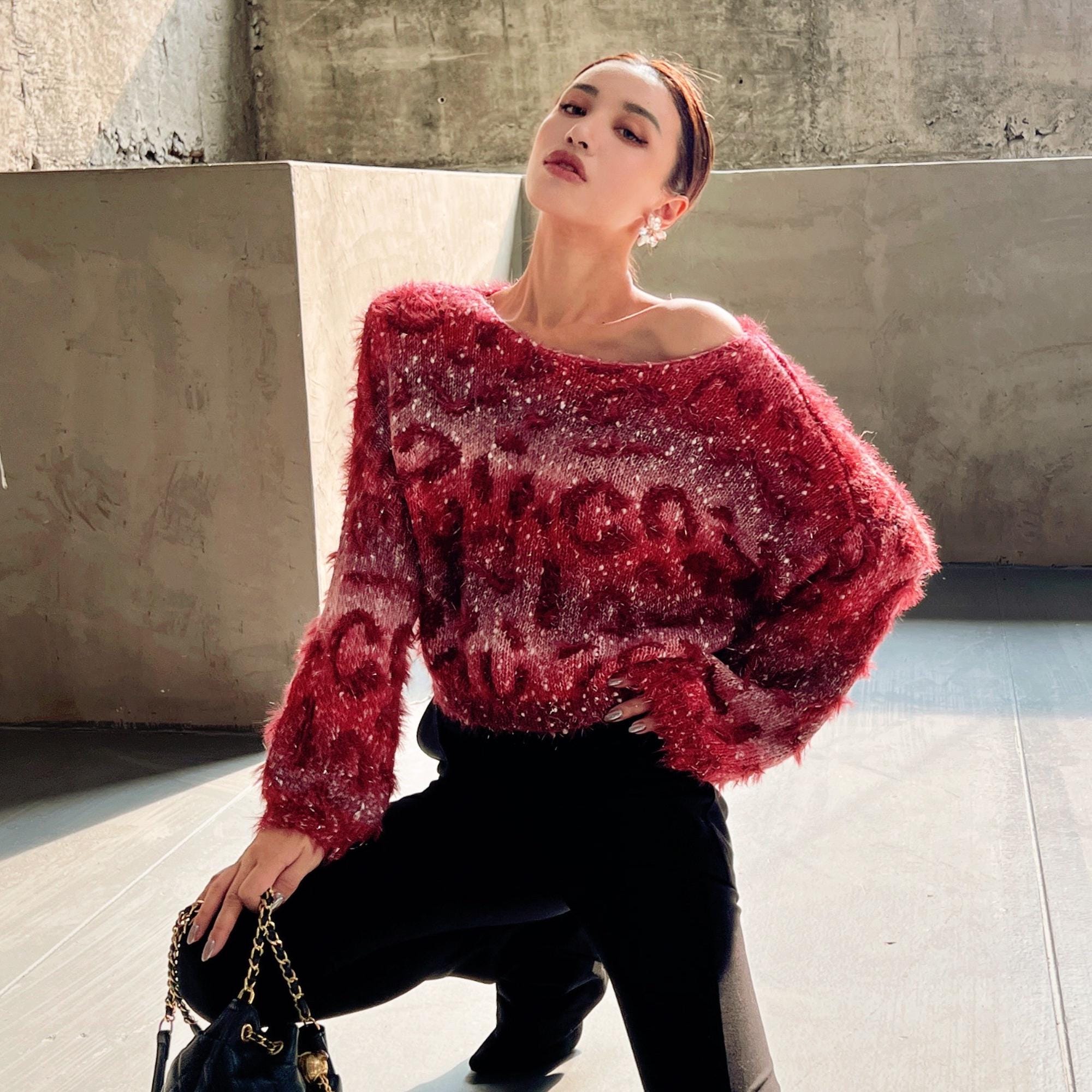A woman wearing a red sweater with a patterned design, posing in an indoor setting with a concrete wall in the background.
