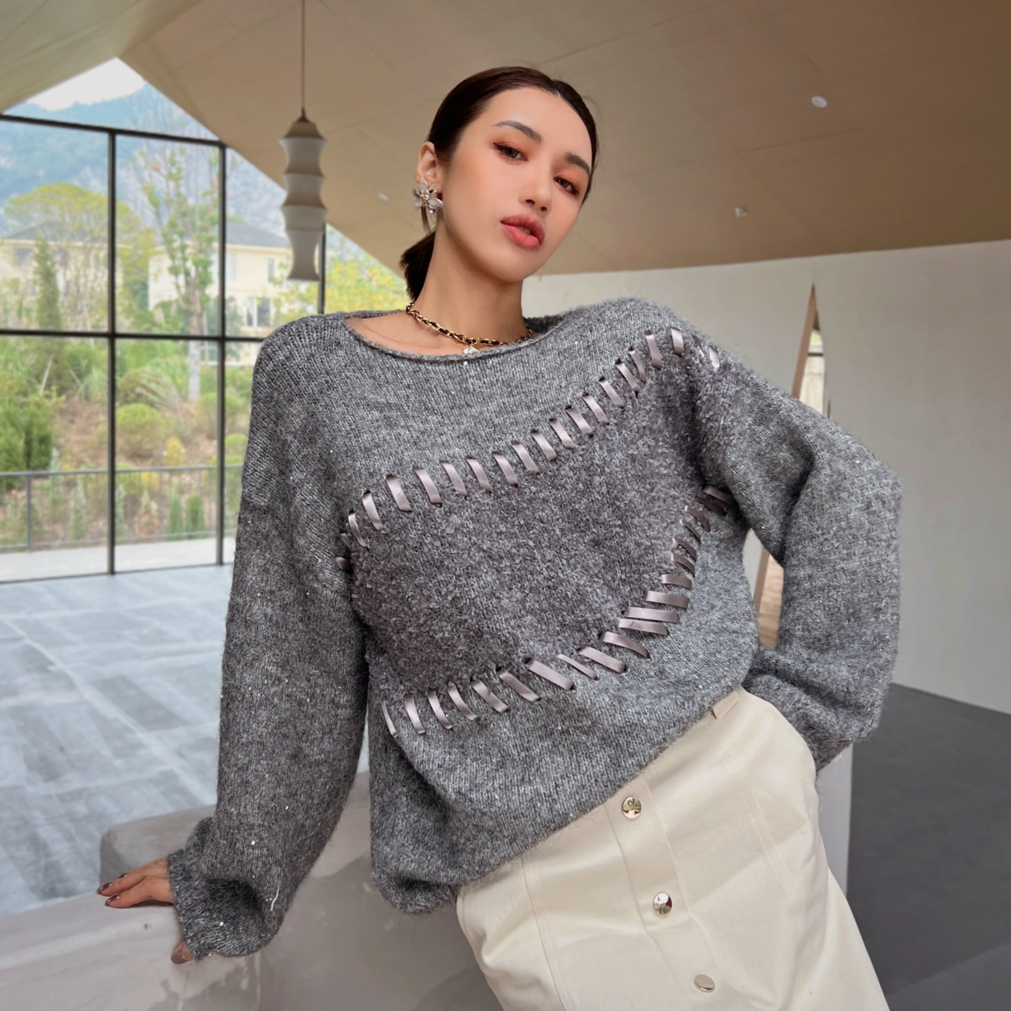 A woman wearing a gray sweater with silver embellishments and a white skirt is posing in a room with large windows overlooking a mountainous landscape.