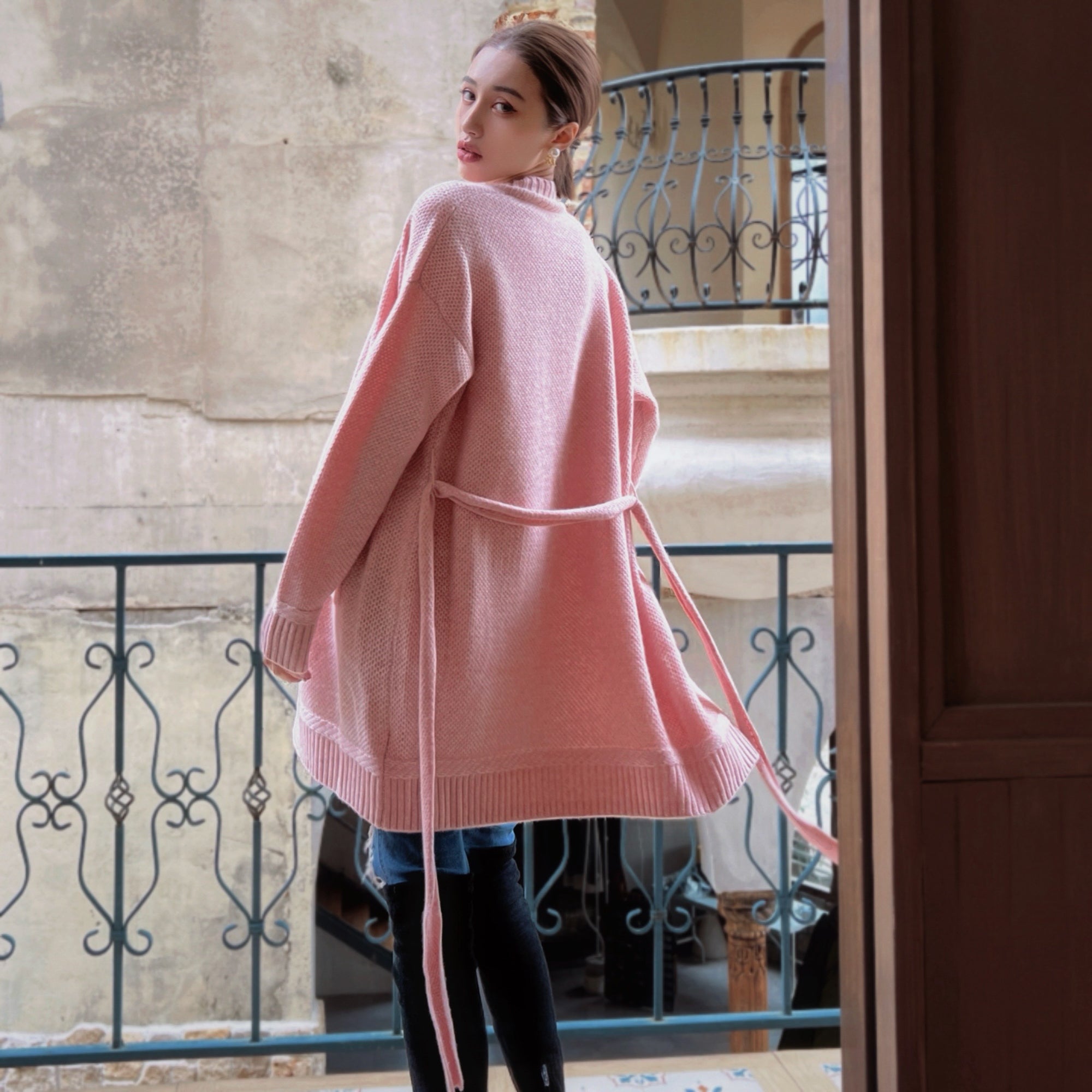 A woman wearing a pink cardigan stands in front of a balcony, looking over her shoulder.