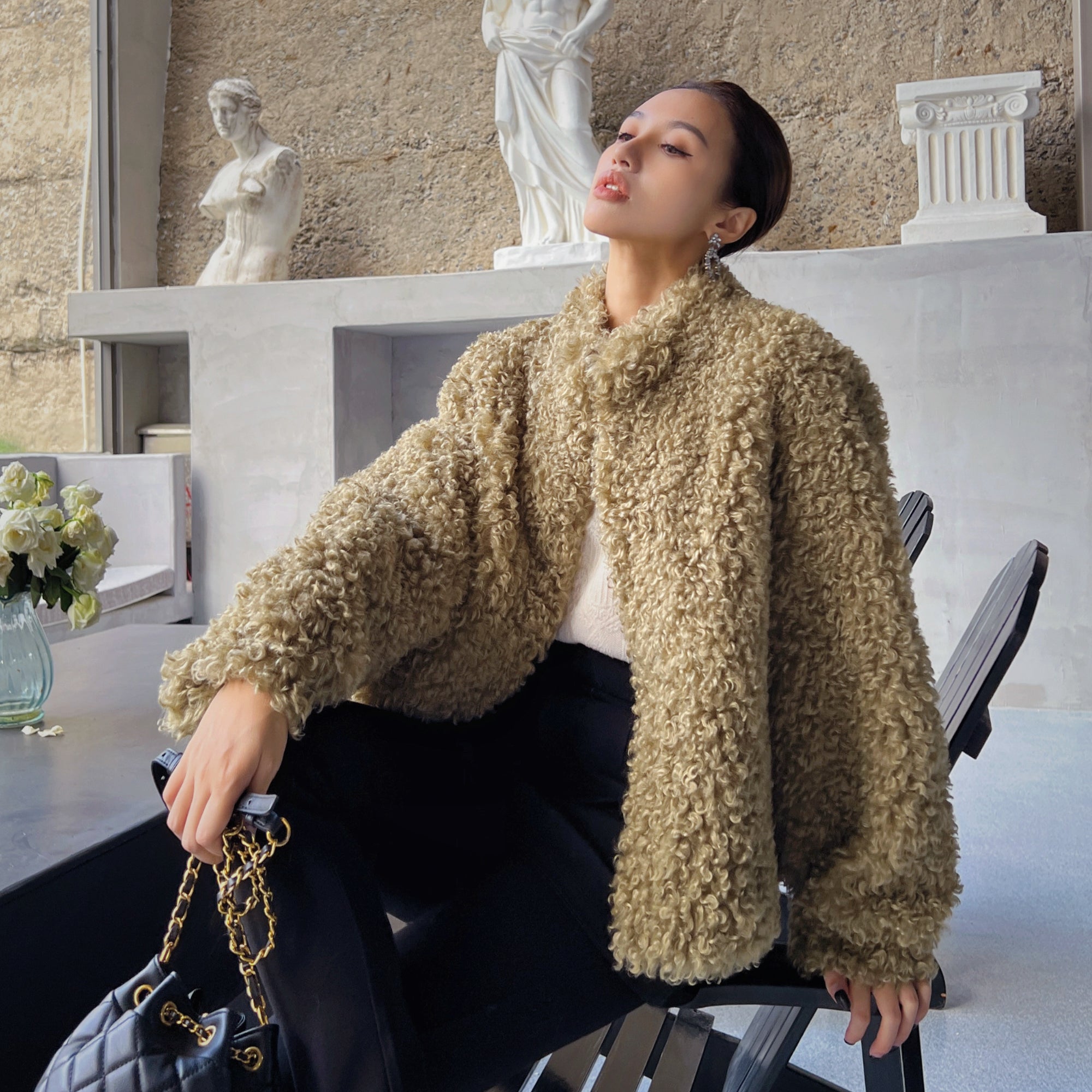 A woman wearing a beige fur coat is sitting on a chair, holding a black purse, and looking up.