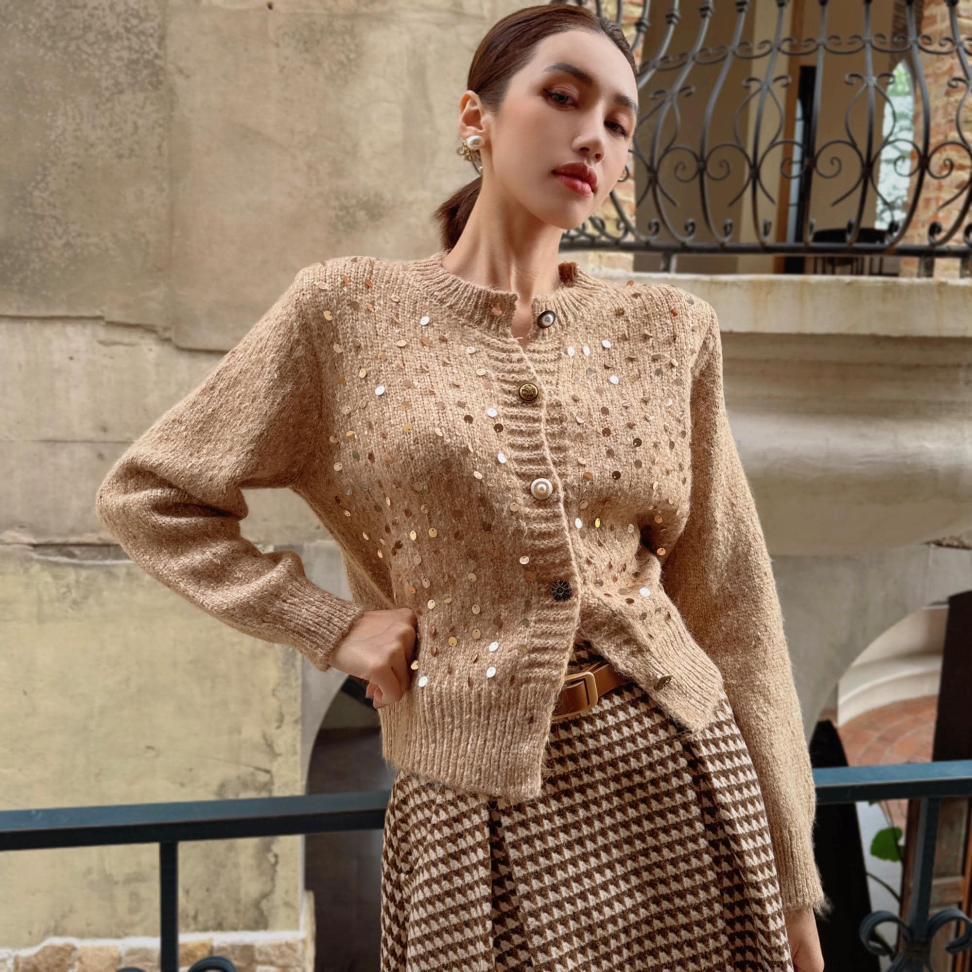 A woman wearing a beige sweater with sparkly buttons and a brown and white patterned skirt, posing on a balcony with a wrought iron railing in the background.