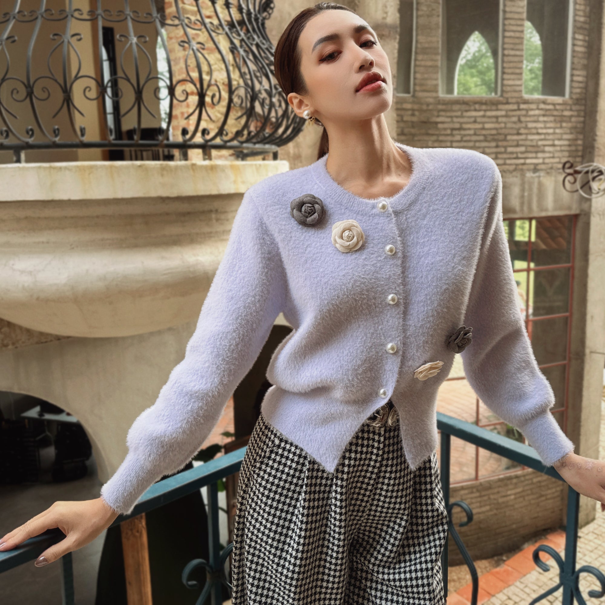 A woman wearing a light purple cardigan with decorative buttons and a black and white checkered skirt stands on a balcony, looking off into the distance.