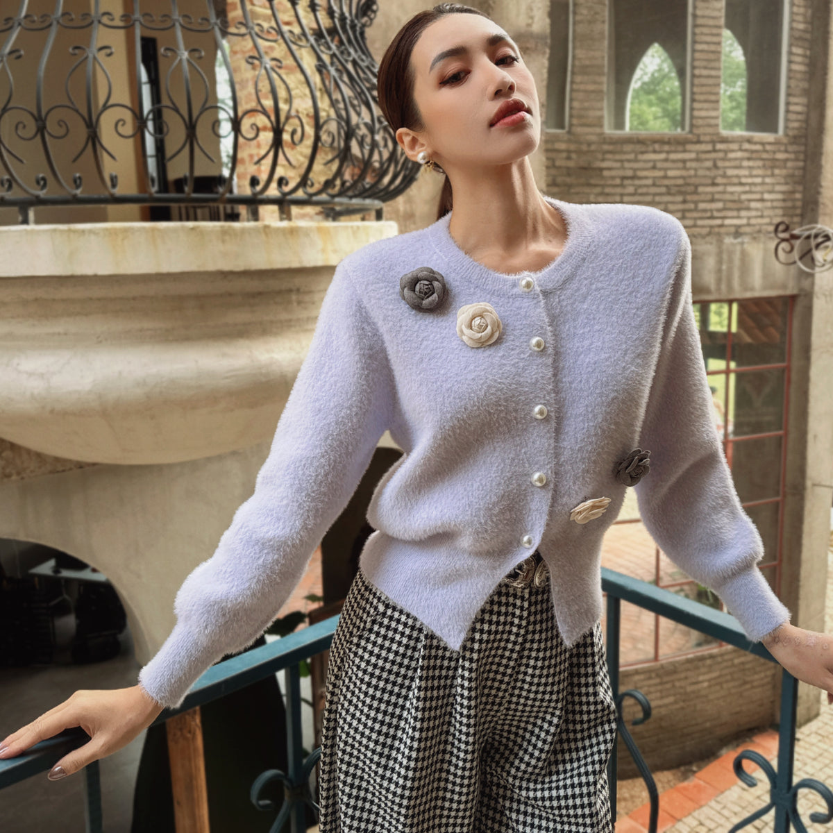 A woman wearing a light purple cardigan with decorative buttons and a black and white checkered skirt stands on a balcony, looking off into the distance.