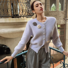 A woman wearing a light purple cardigan with decorative buttons and a black and white checkered skirt stands on a balcony, looking off into the distance.