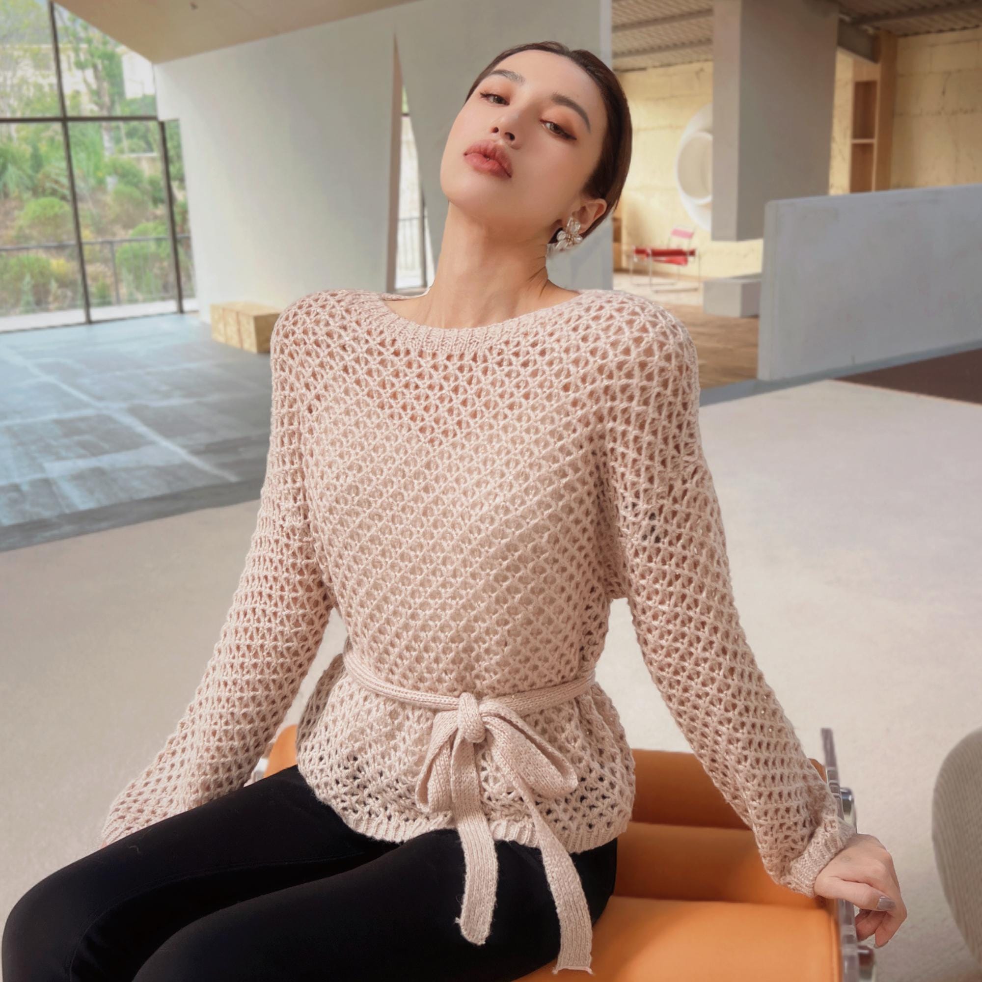 A woman wearing a beige knitted sweater with a bow detail, sitting on an orange bench in an indoor setting.