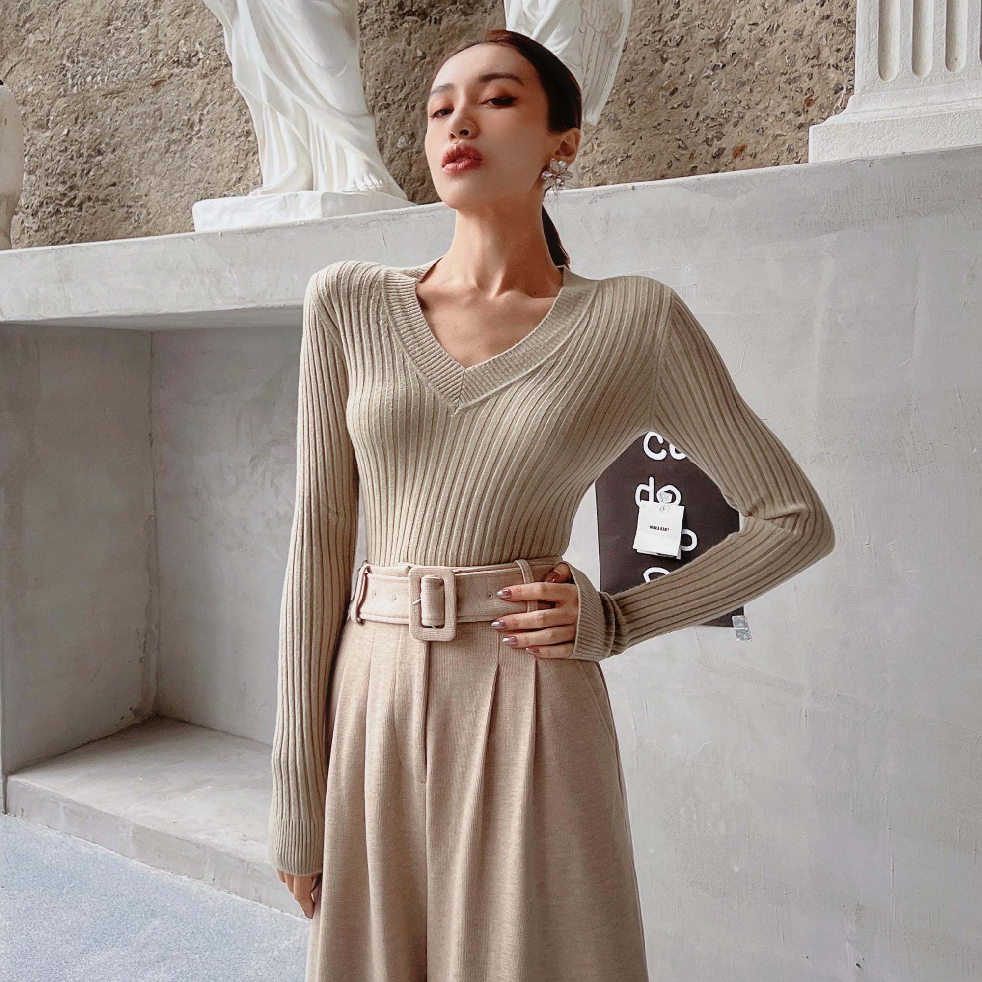 A woman wearing a beige sweater and a beige skirt stands in front of a white wall, with a statue visible in the background.