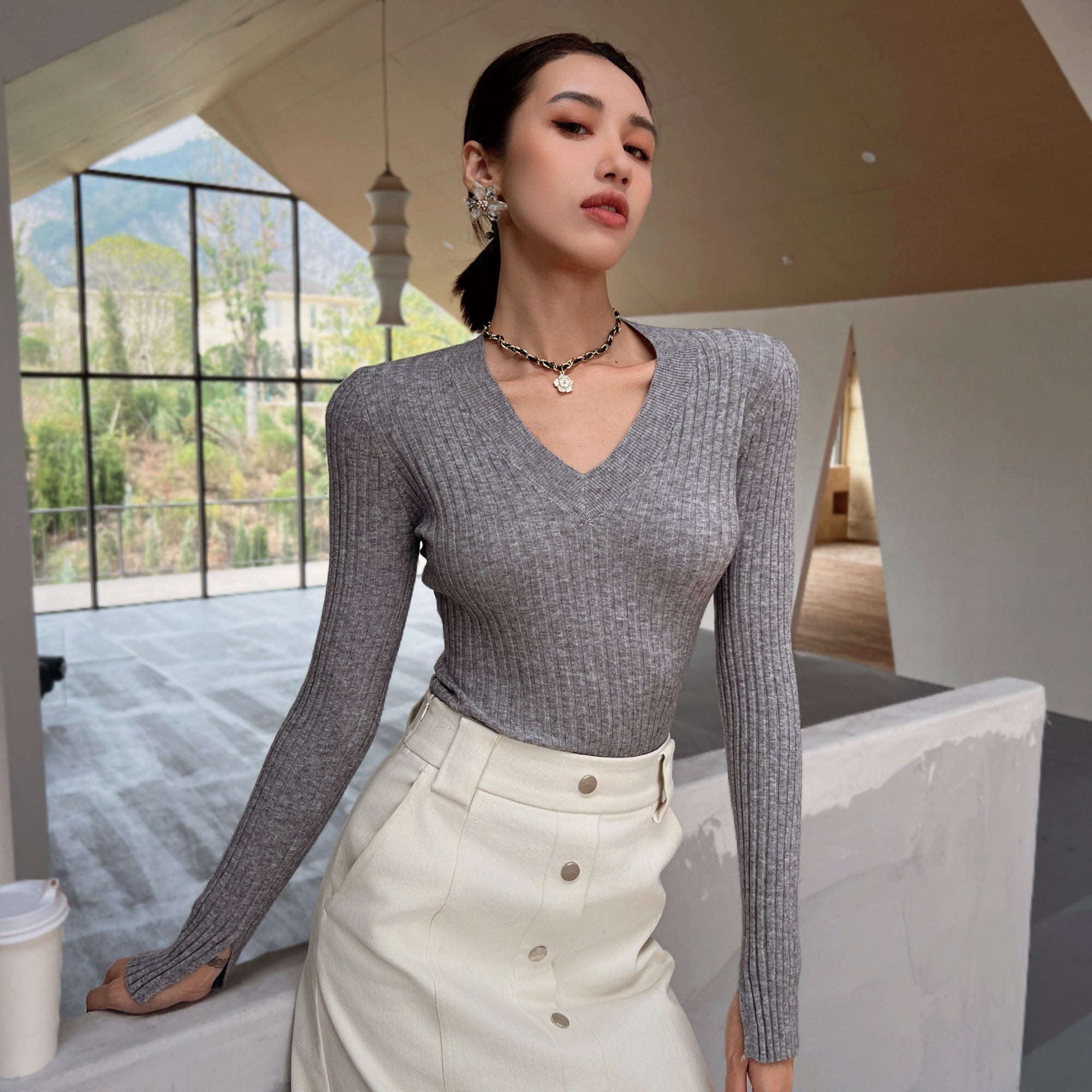 A woman wearing a gray sweater and a white skirt stands in a room with a large window overlooking a mountainous landscape.