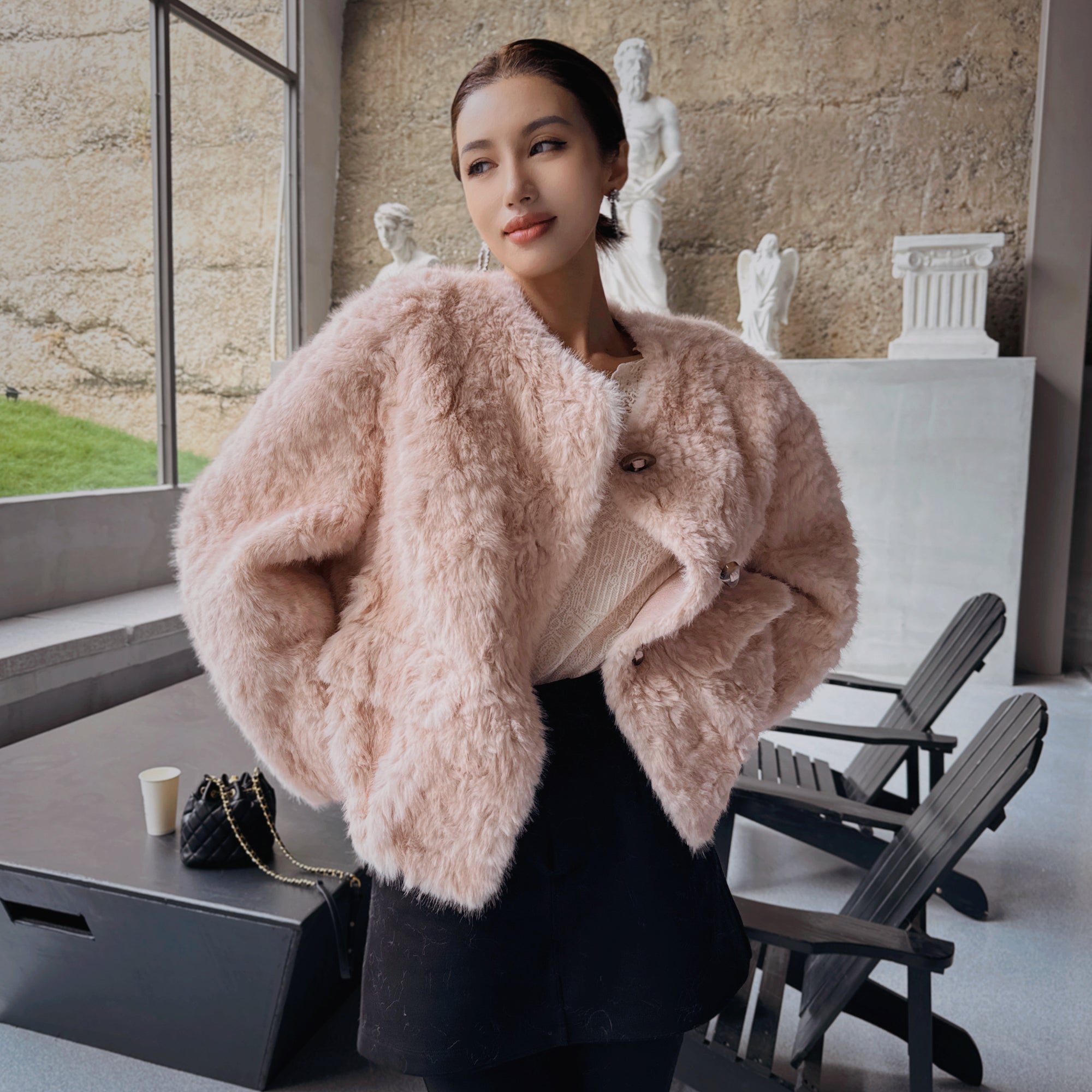 A woman wearing a pink fur coat stands in a room with a window, a statue, and a chair.