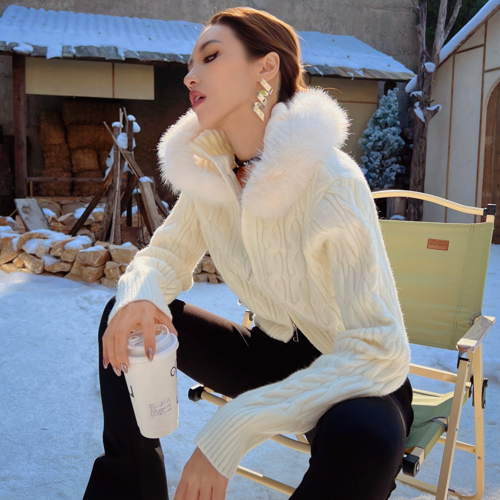 A woman in a white sweater with a fur collar sits on a chair, holding a white cup, with a snowy background and a building with a tiled roof.