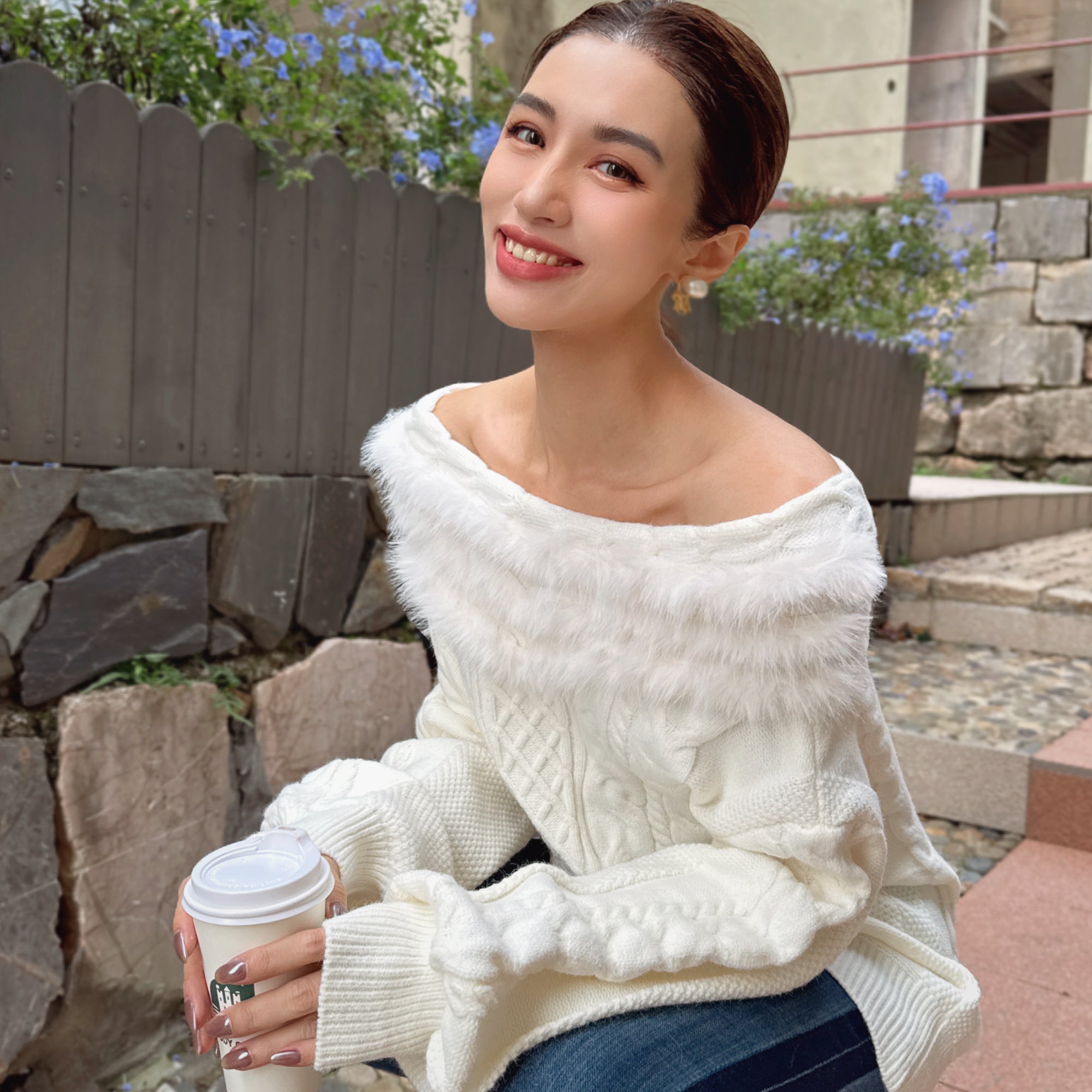 A young woman in a white sweater is sitting on a stone bench, holding a coffee cup and smiling at the camera.