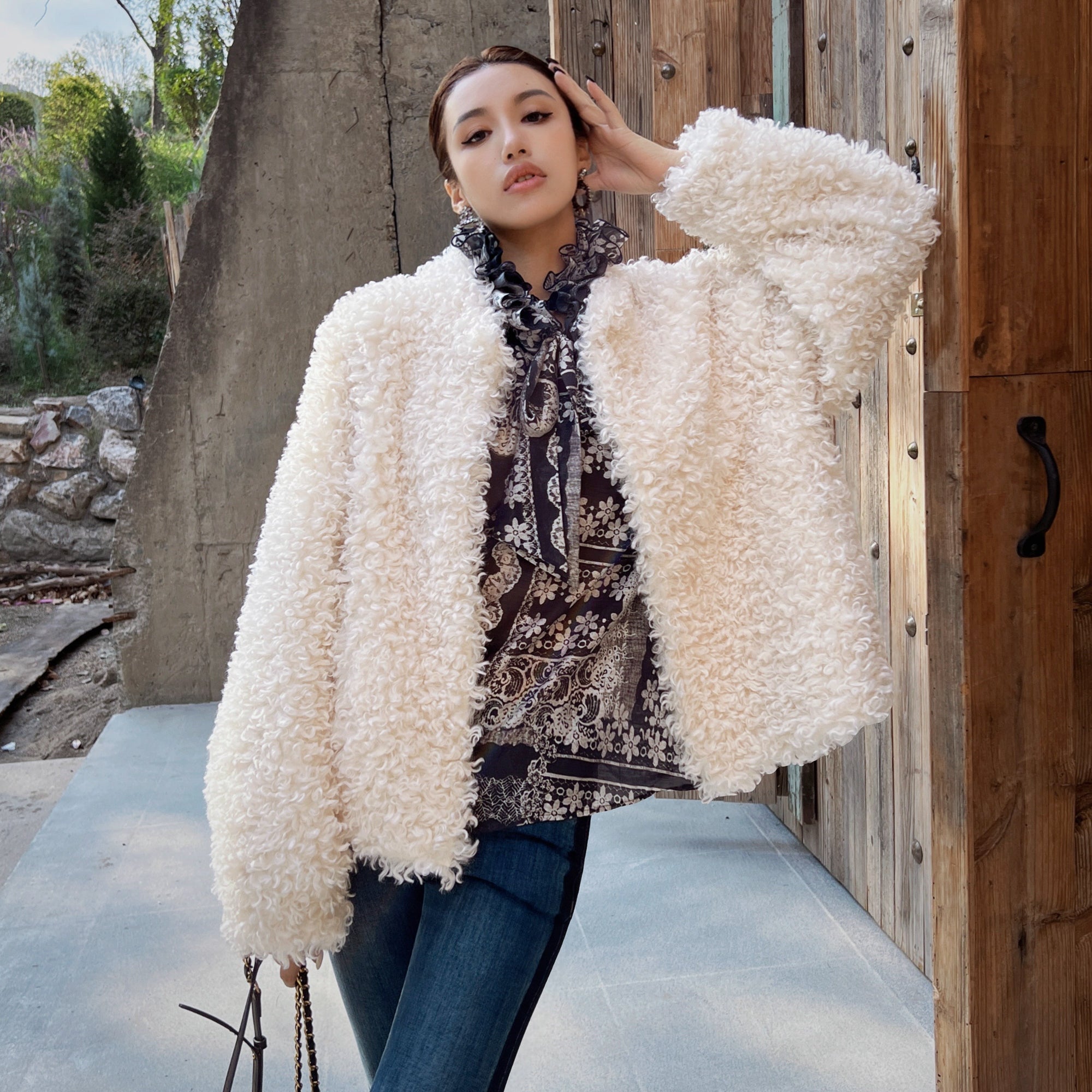 A woman wearing a white fluffy coat and holding a purse stands in front of a wooden structure.
