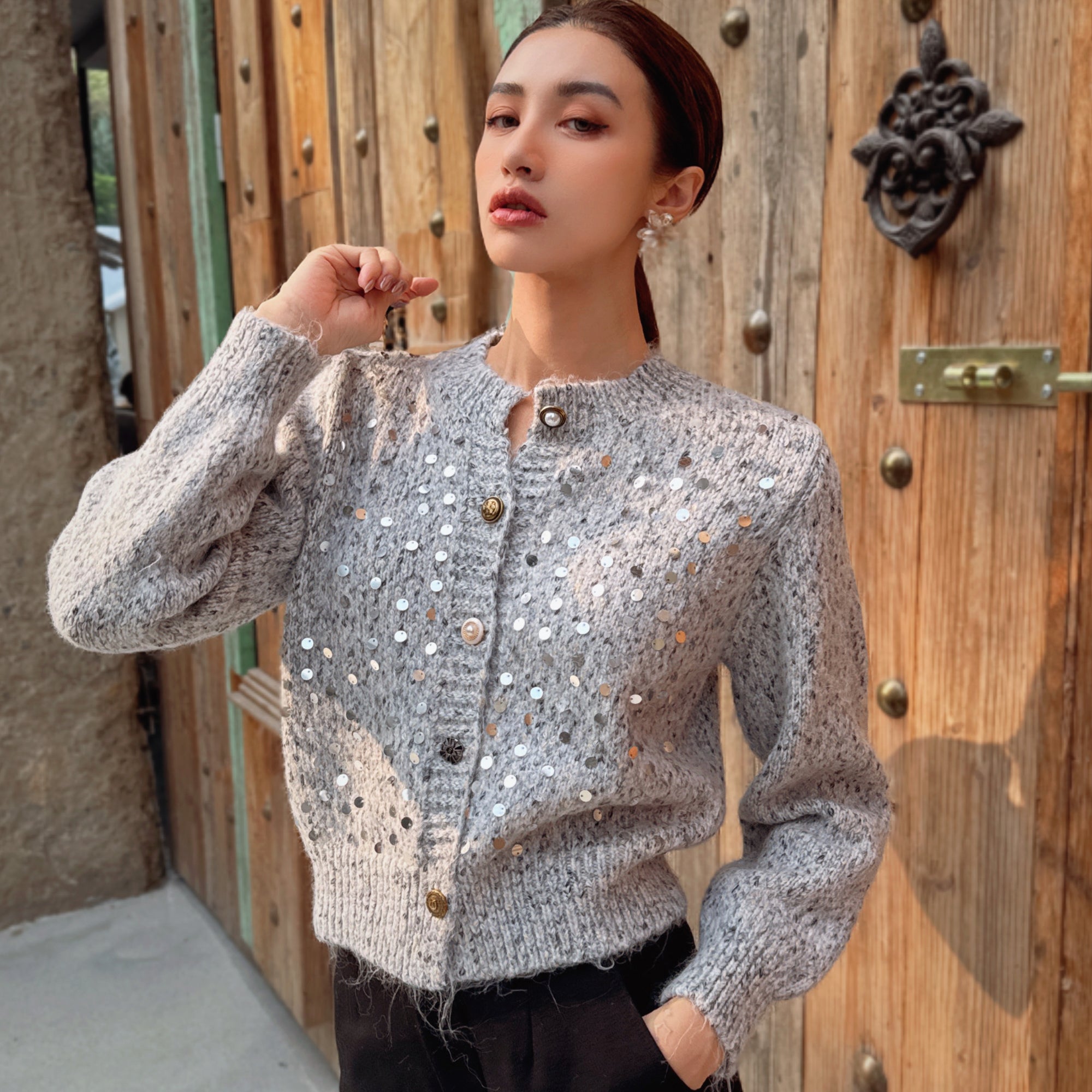 A woman wearing a gray sweater with silver buttons and a floral pattern stands in front of a wooden door.
