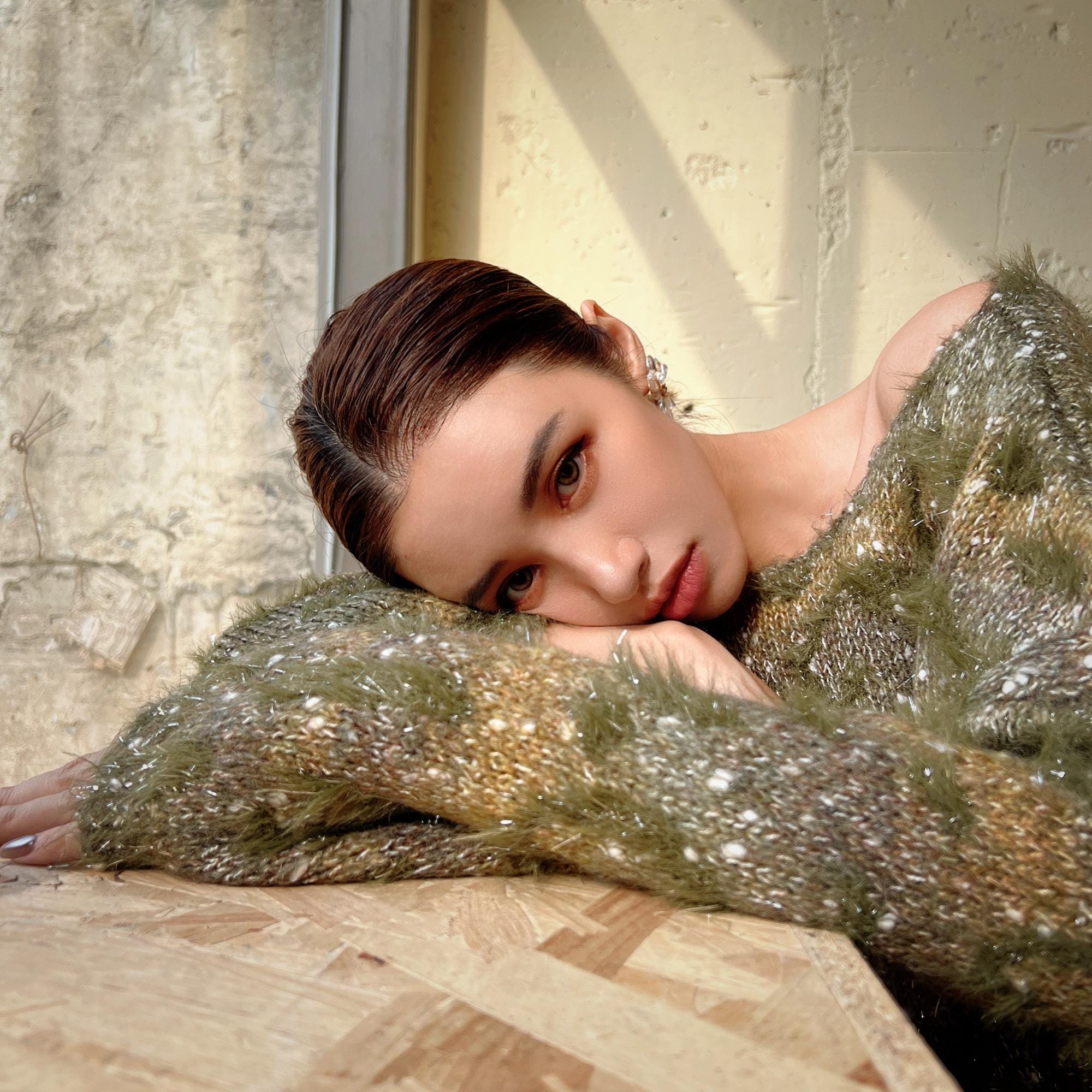 A woman with brown hair is resting her head on her arm, wearing a green and gold patterned sweater, in a room with a concrete wall and window.
