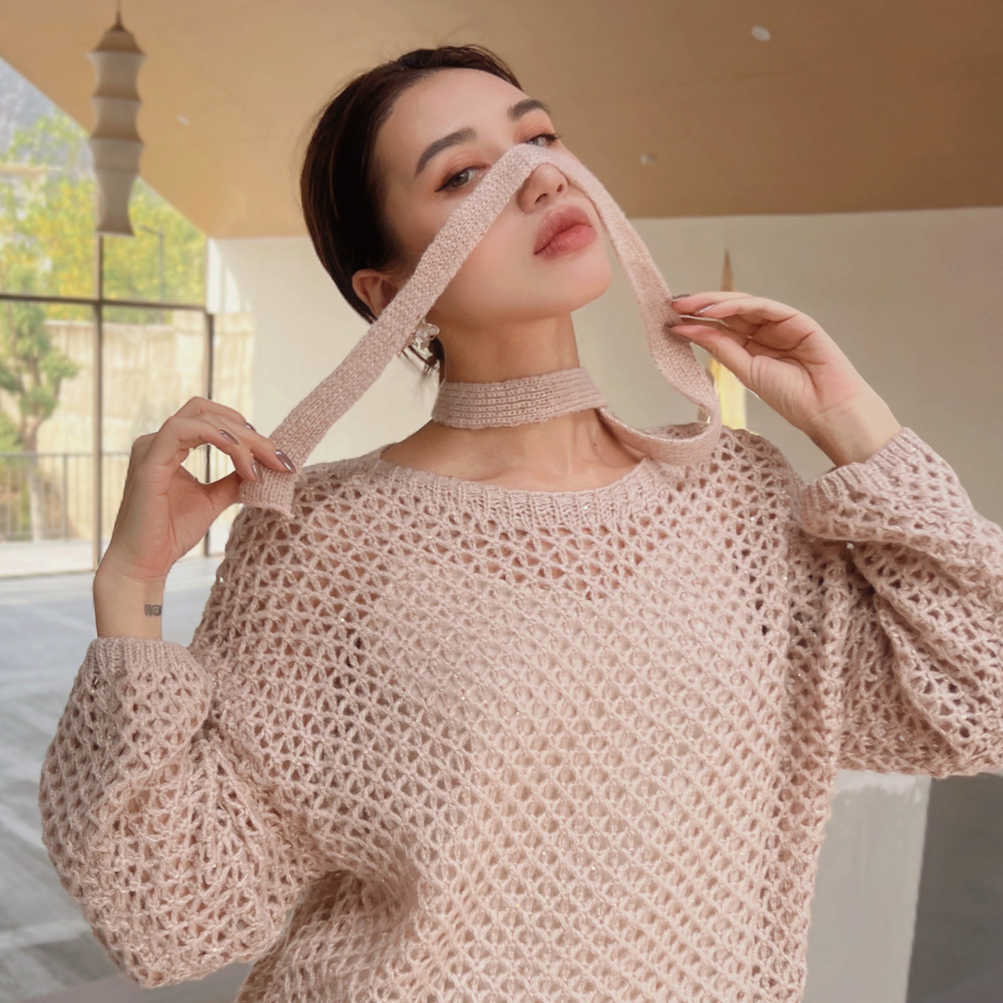 A woman wearing a pink knitted sweater and holding a scarf to her face, posing in an indoor setting.