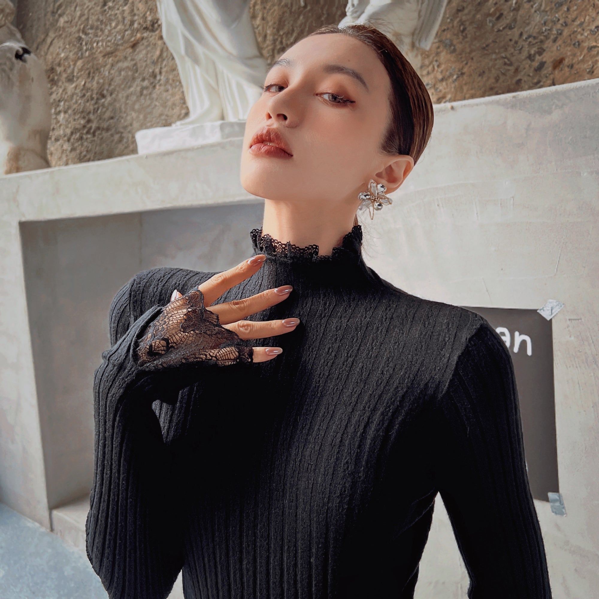 A woman wearing a black sweater and a black lace glove poses for a portrait against a stone wall.