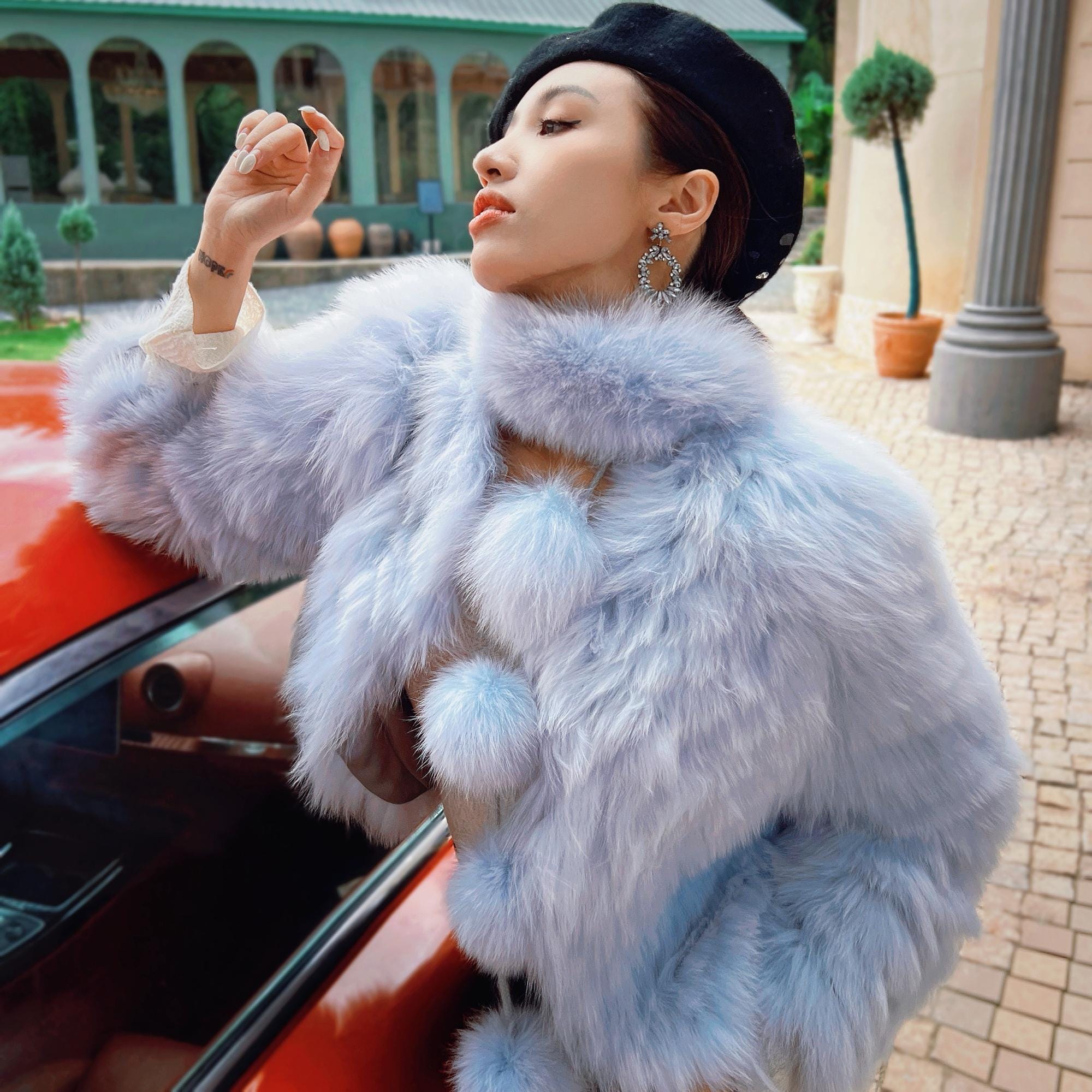 A woman in a blue fur coat and black hat stands next to a red car, with a building and potted plants in the background.