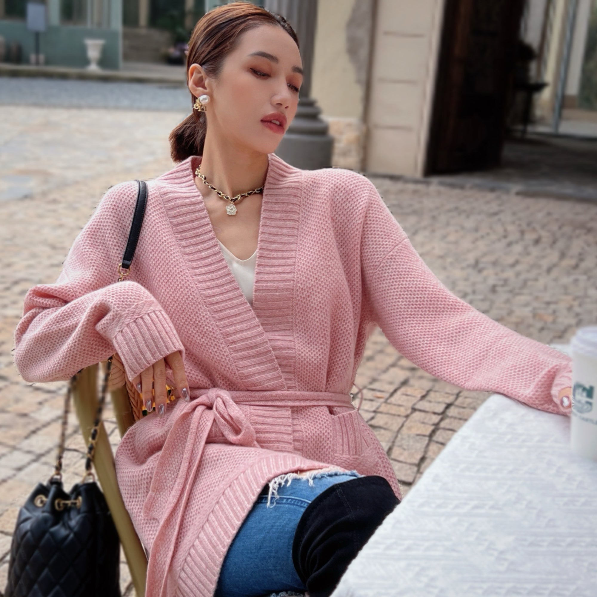 A woman wearing a pink cardigan and jeans is sitting at a table outdoors, with a handbag next to her.