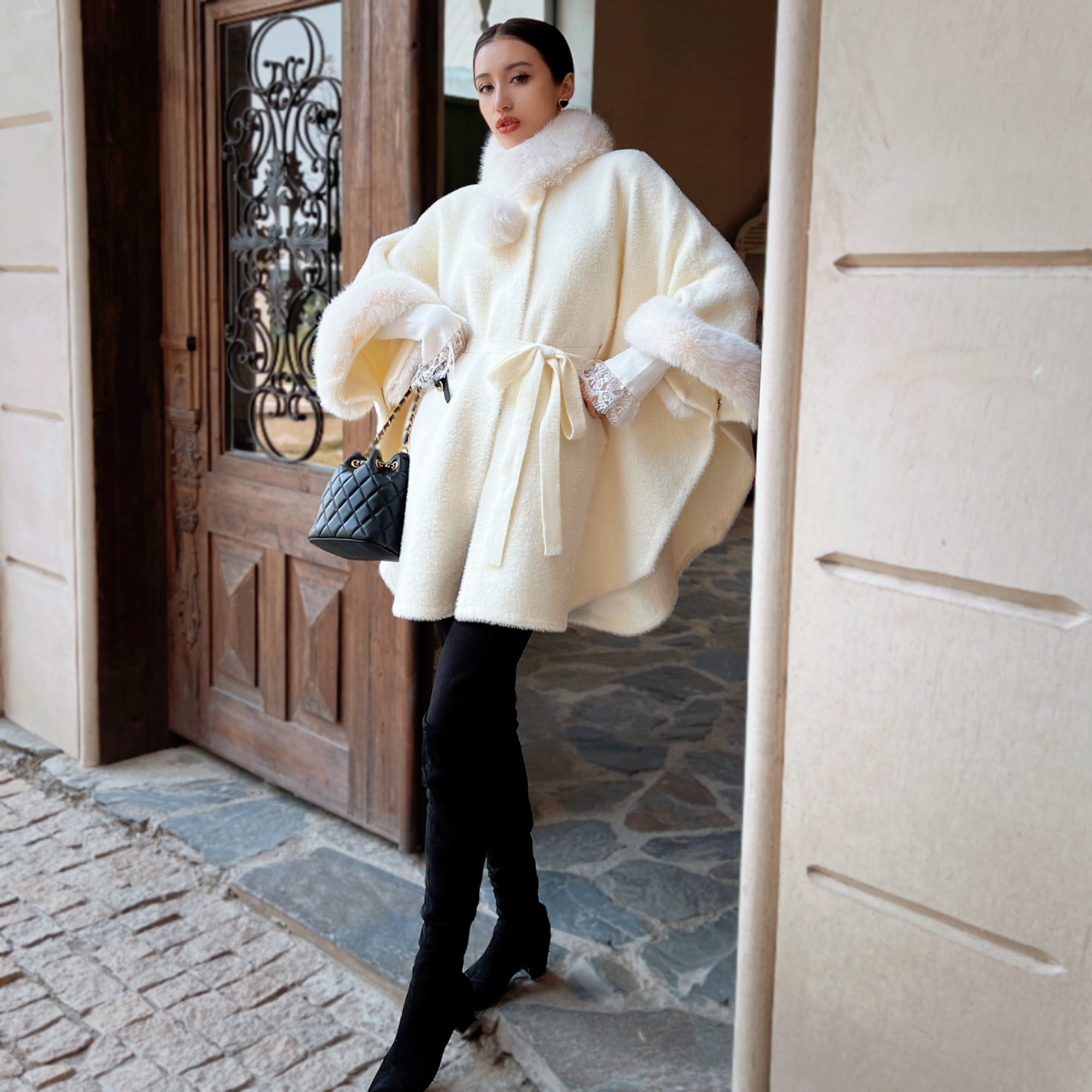 A woman in a white fur-trimmed coat stands in front of a wooden door, holding a black handbag.