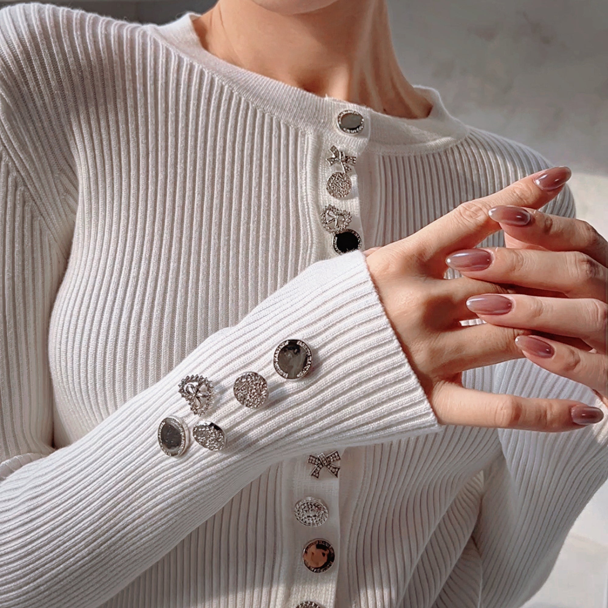 A close-up of a person wearing a white sweater with several silver buttons and rhinestones or crystals attached to the sleeves.