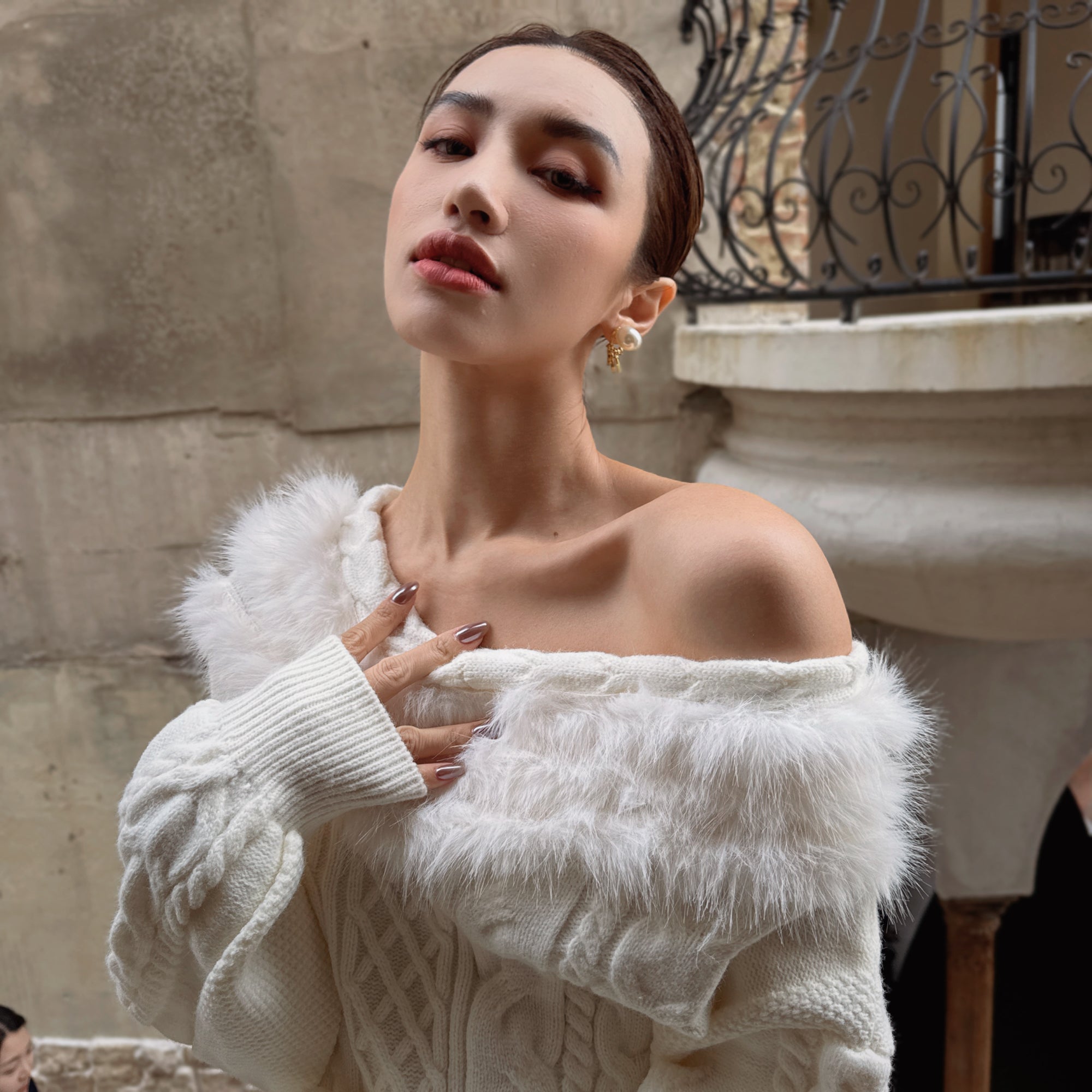 A woman wearing a white fur coat and posing with her hand on her chest against a stone wall background.