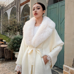 A woman wearing a white fur coat with a fur collar stands in front of a building with arched windows.