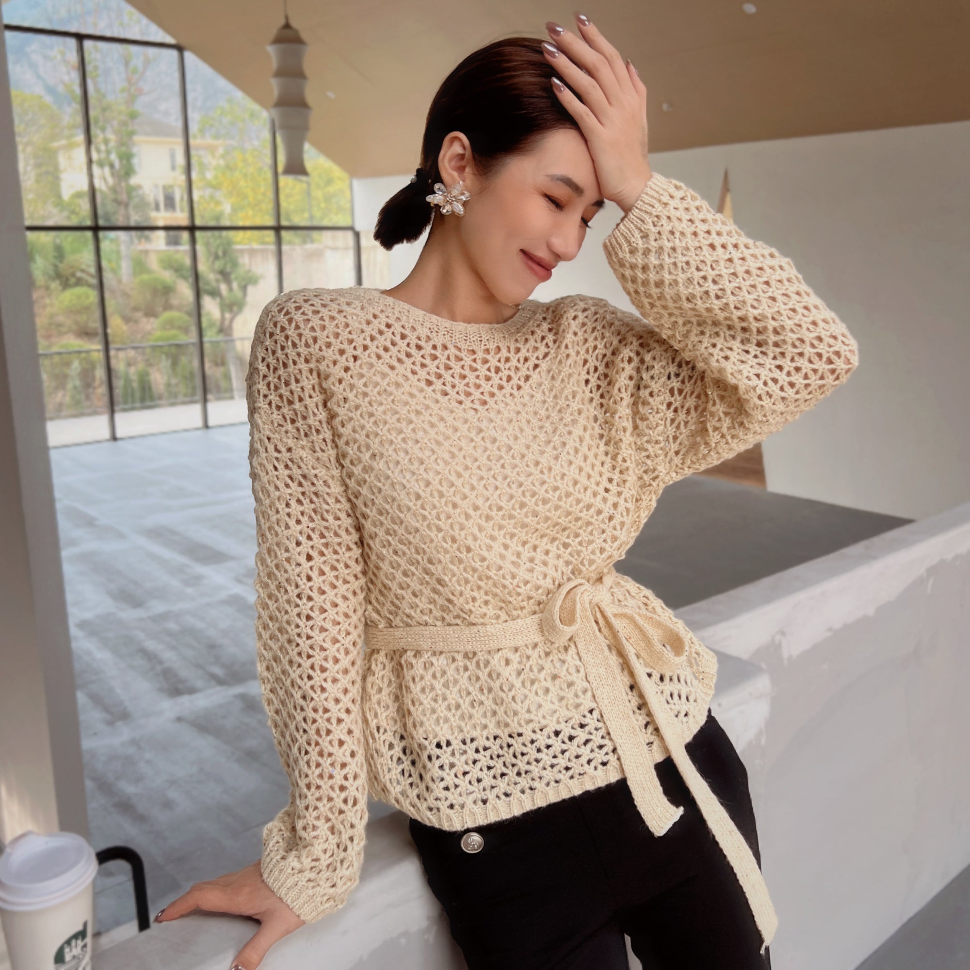 A woman wearing a beige crocheted sweater with a tie, posing with her hand on her head in a relaxed manner.