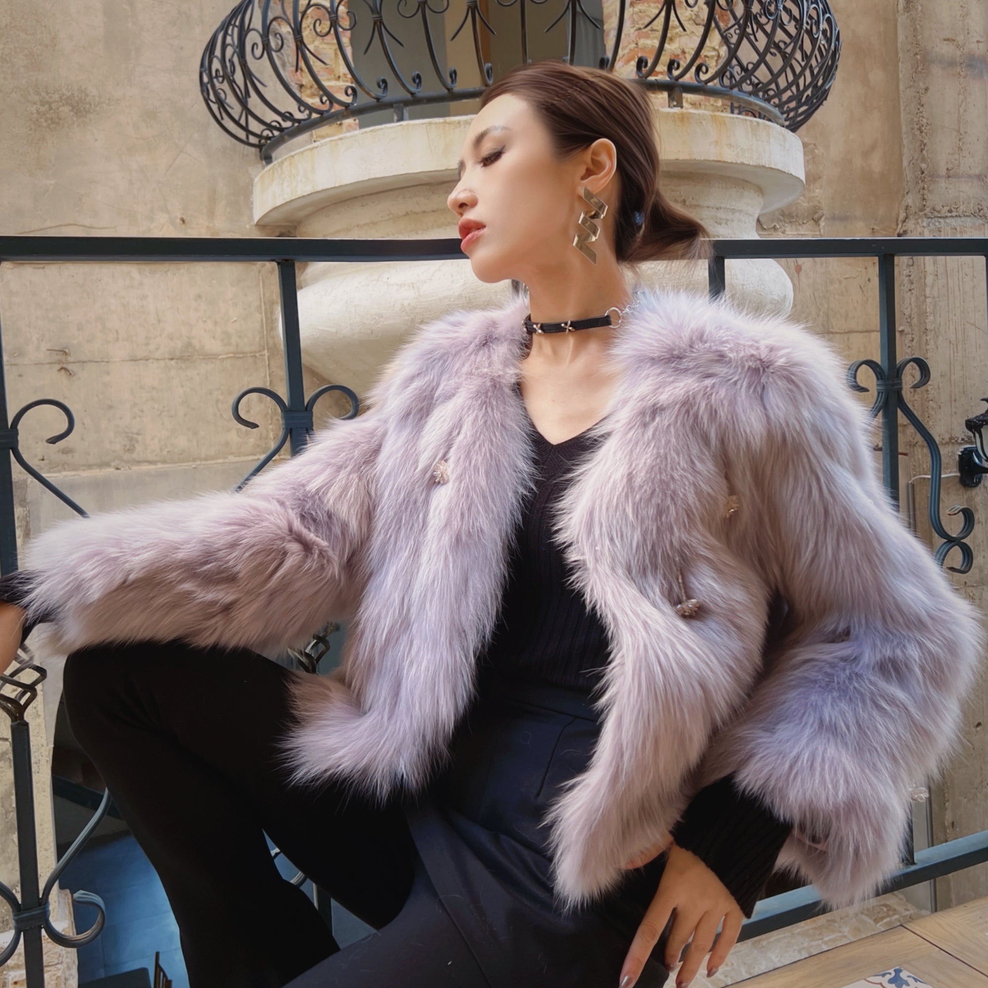 A woman wearing a fur coat and black outfit is posing in front of a wrought iron fence.