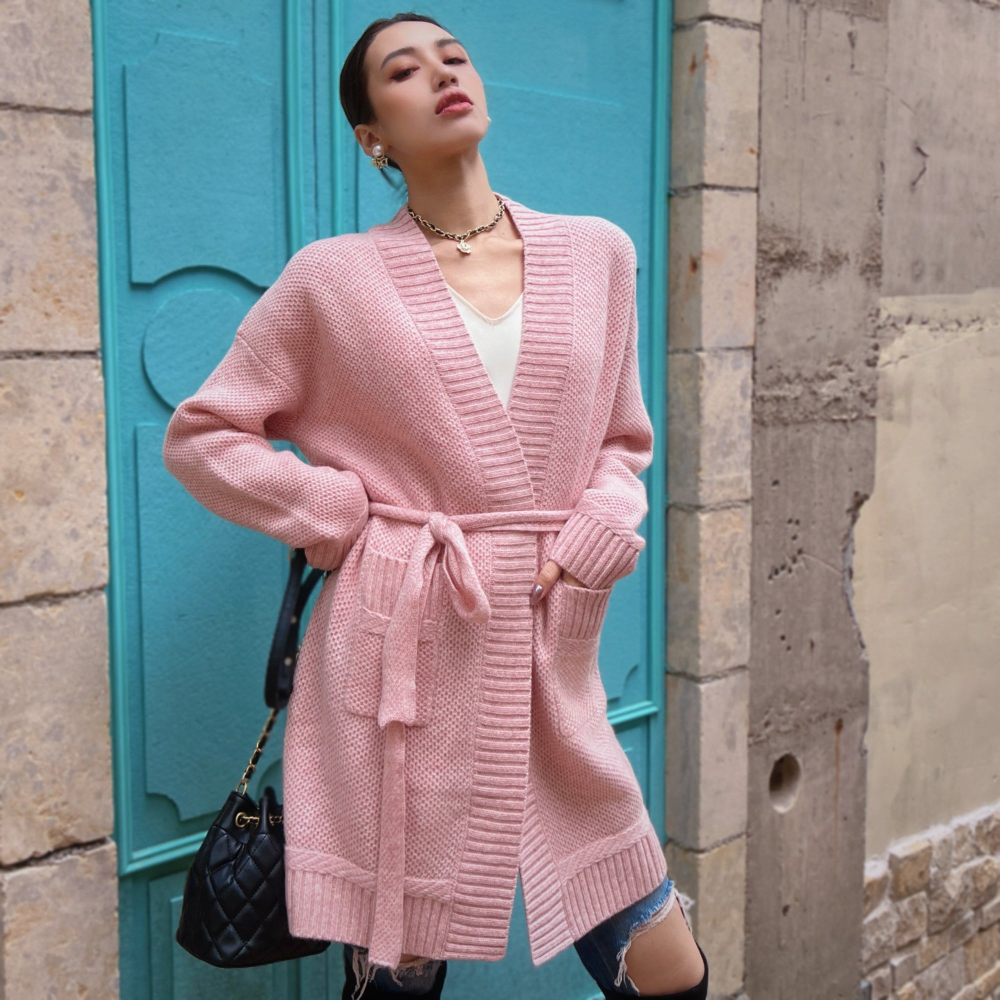 A woman wearing a pink cardigan stands in front of a blue door, with her hands in her pockets and a black purse hanging from her shoulder.