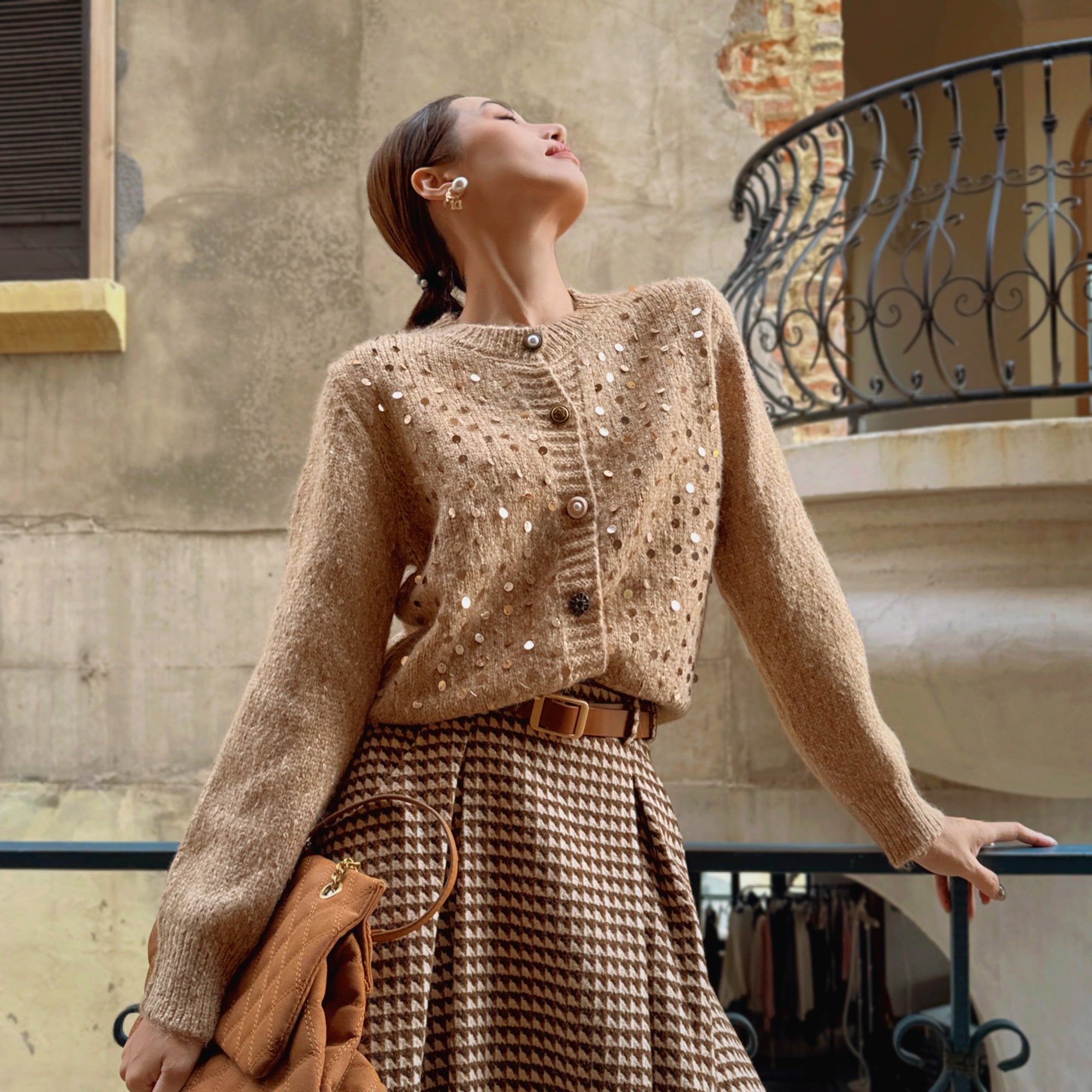 A woman wearing a beige sweater with white polka dots and a brown skirt, standing on a balcony with a wrought iron railing.