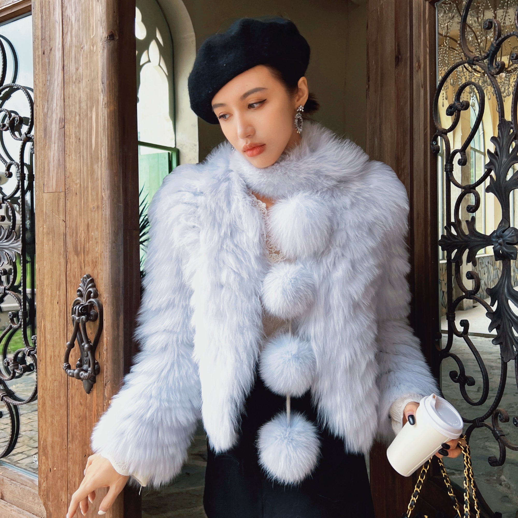 A woman wearing a white fur coat and a black hat stands in front of a wooden door, holding a white coffee cup.