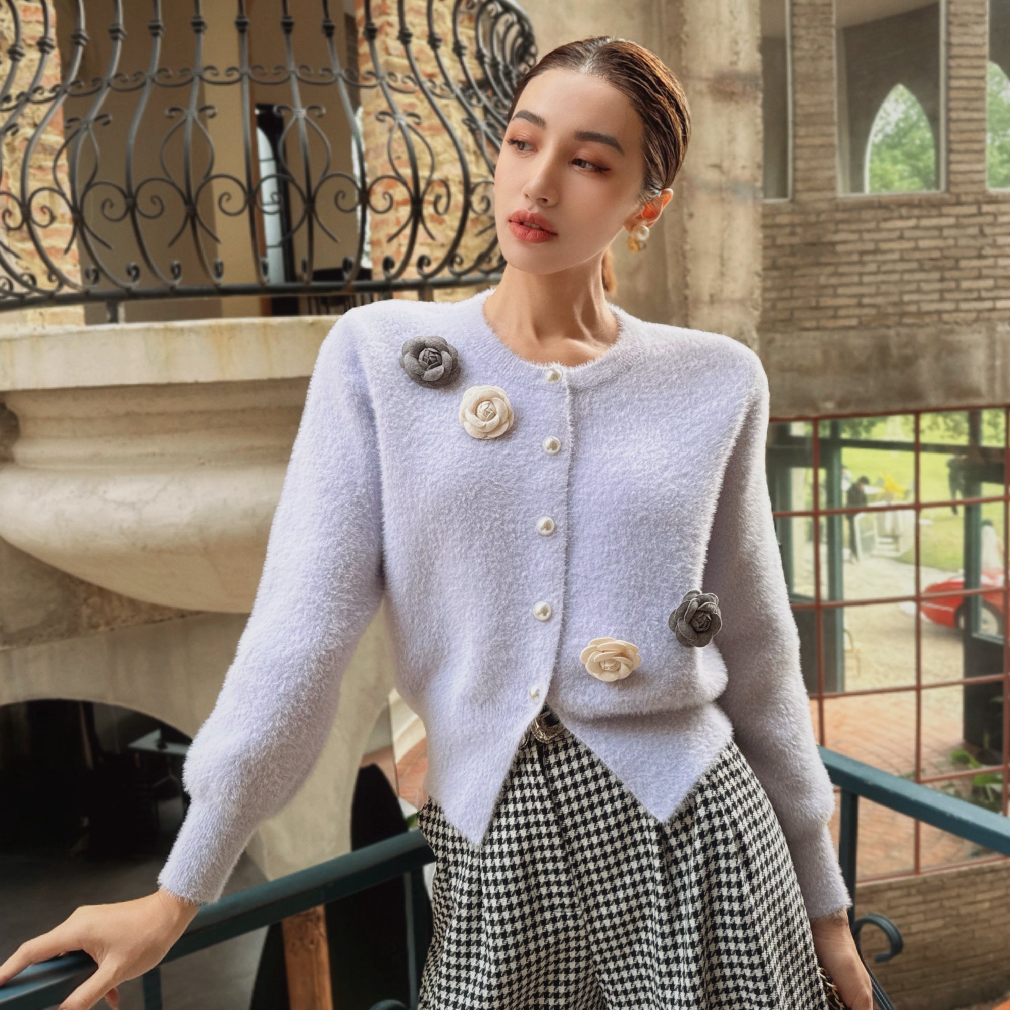 A woman wearing a light purple cardigan with floral embellishments and a black and white checkered skirt, posing on a balcony with a wrought iron railing and a brick building in the background.