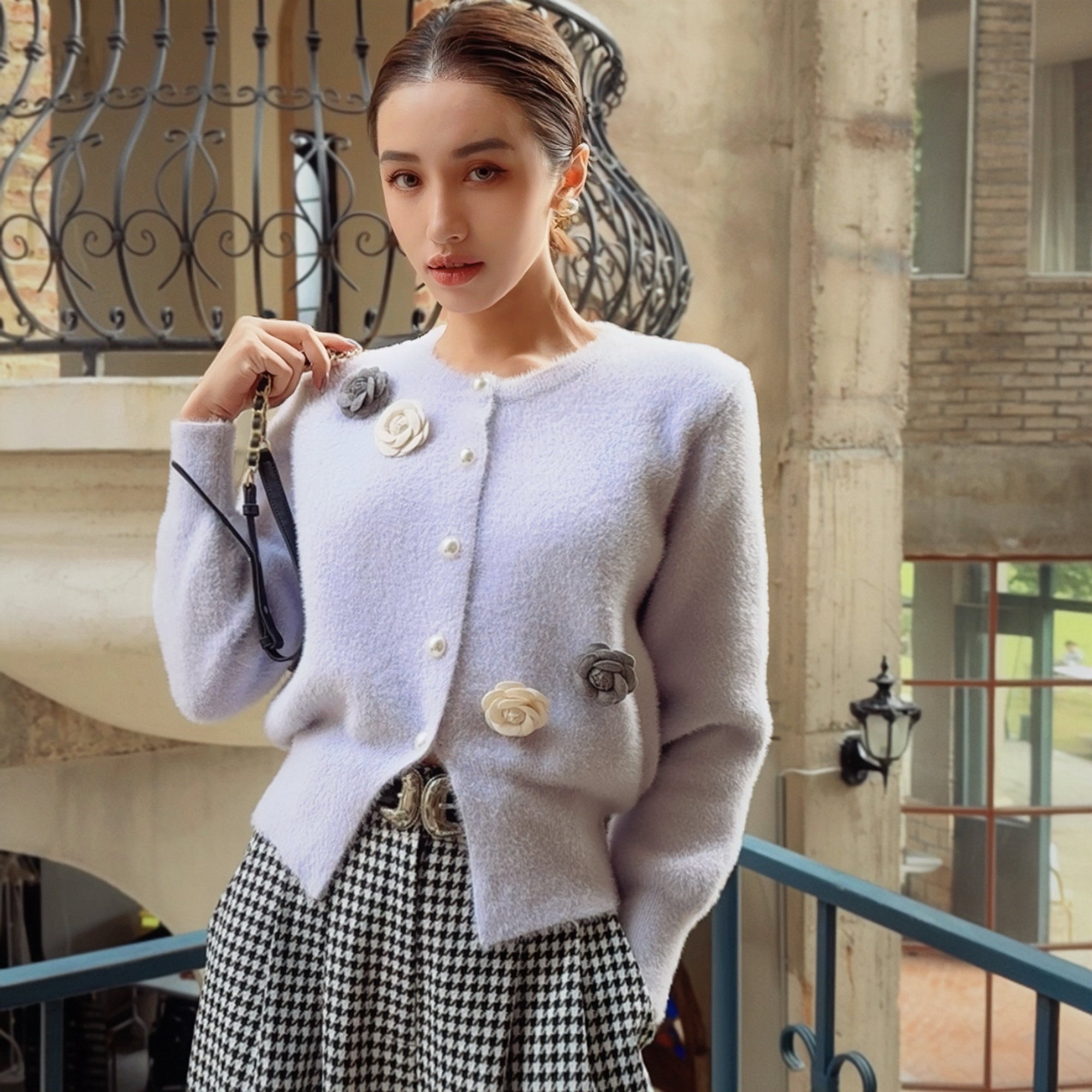 A woman wearing a white cardigan with floral embellishments, posing on a balcony with a black wrought iron railing in the background.