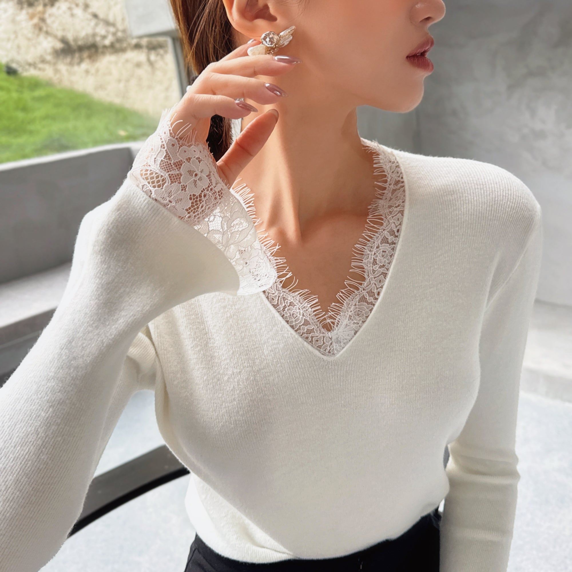 A woman wearing a white lace sweater with a v-neck and long sleeves, posing with her hand on her chin.