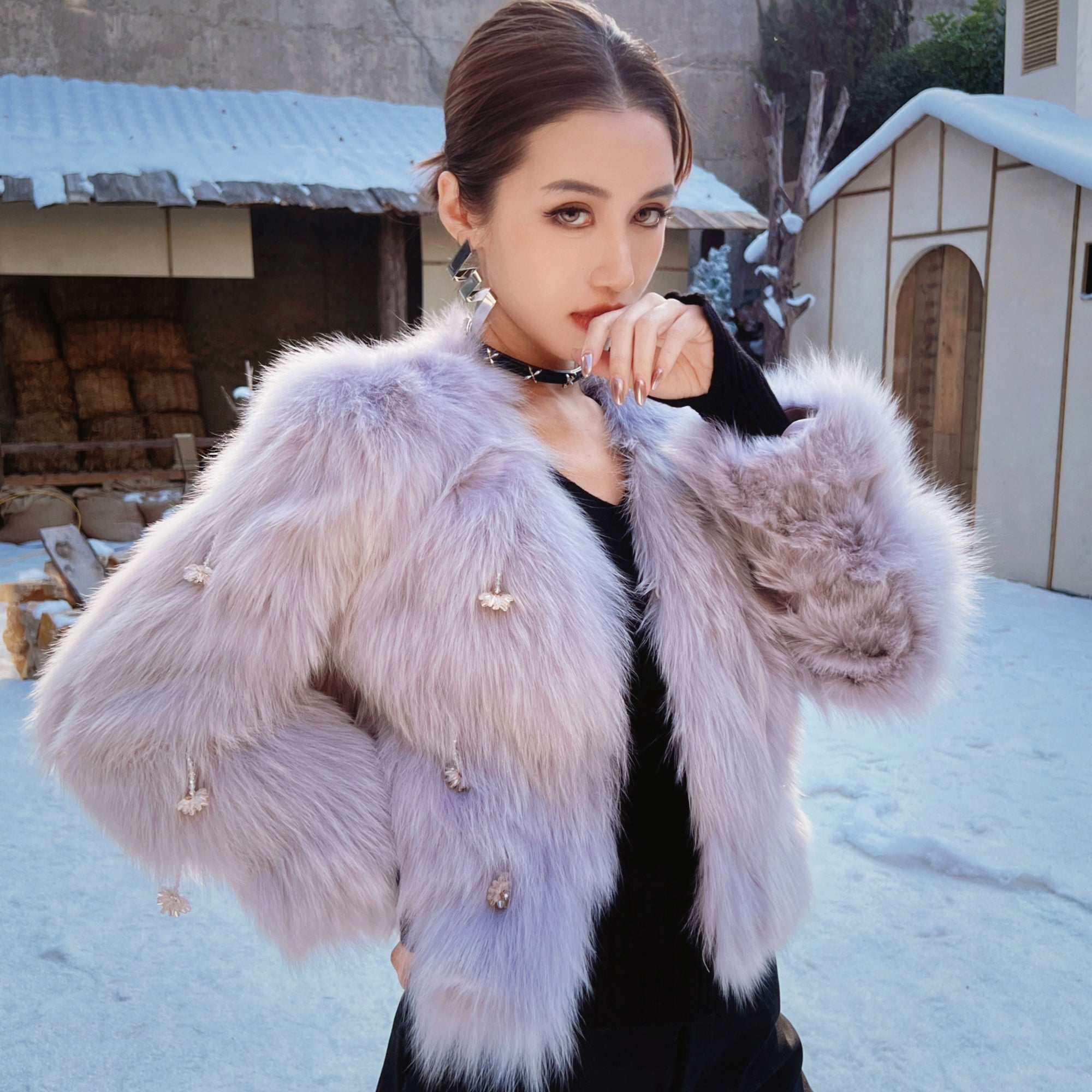 A woman wearing a fur coat stands in front of a snowy building, with her hand on her chin.
