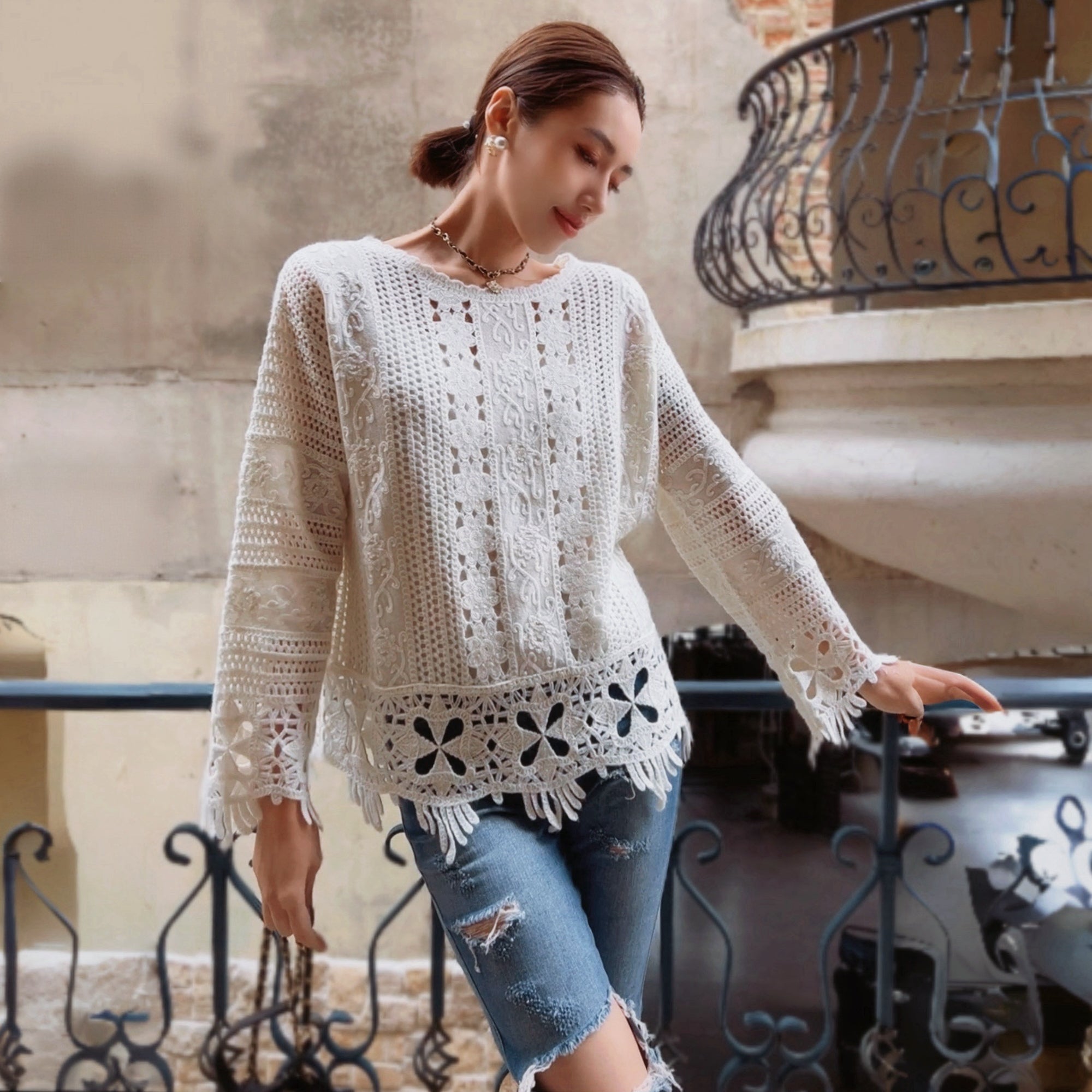 A woman wearing a white lace top and distressed jeans stands in front of a wrought iron railing, with a stone wall and a balcony visible in the background.