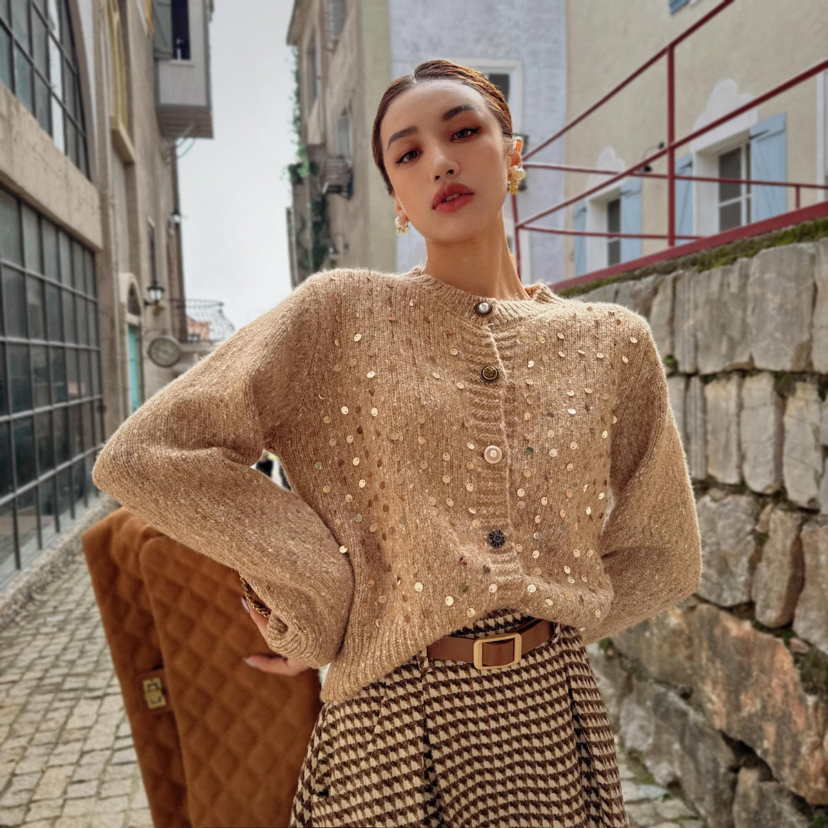 A woman wearing a beige sweater with white polka dots and a brown skirt stands in front of a stone wall.