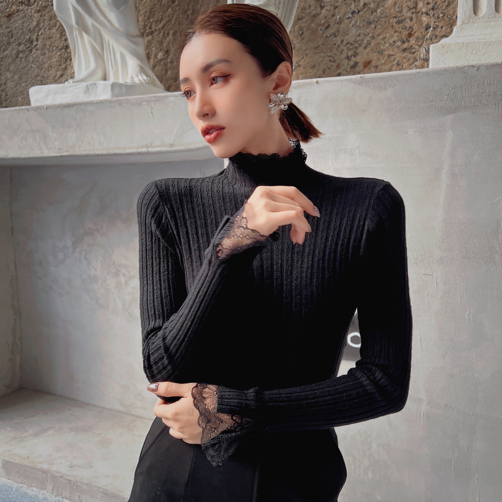 A woman wearing a black sweater with lace sleeves stands in front of a stone wall, looking off to the side.