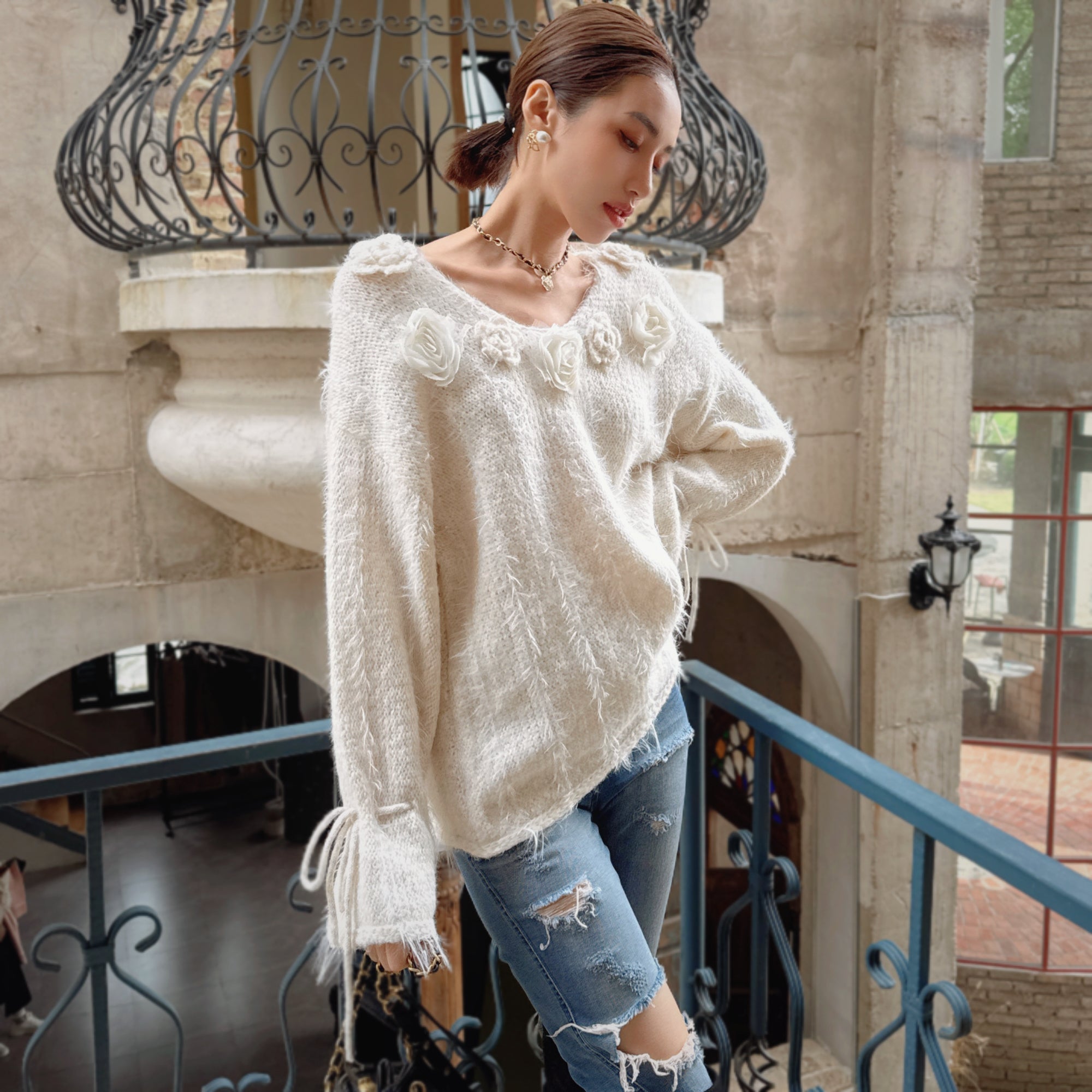 A woman wearing a white sweater with flower designs and ripped jeans stands on a balcony, looking down at the ground.