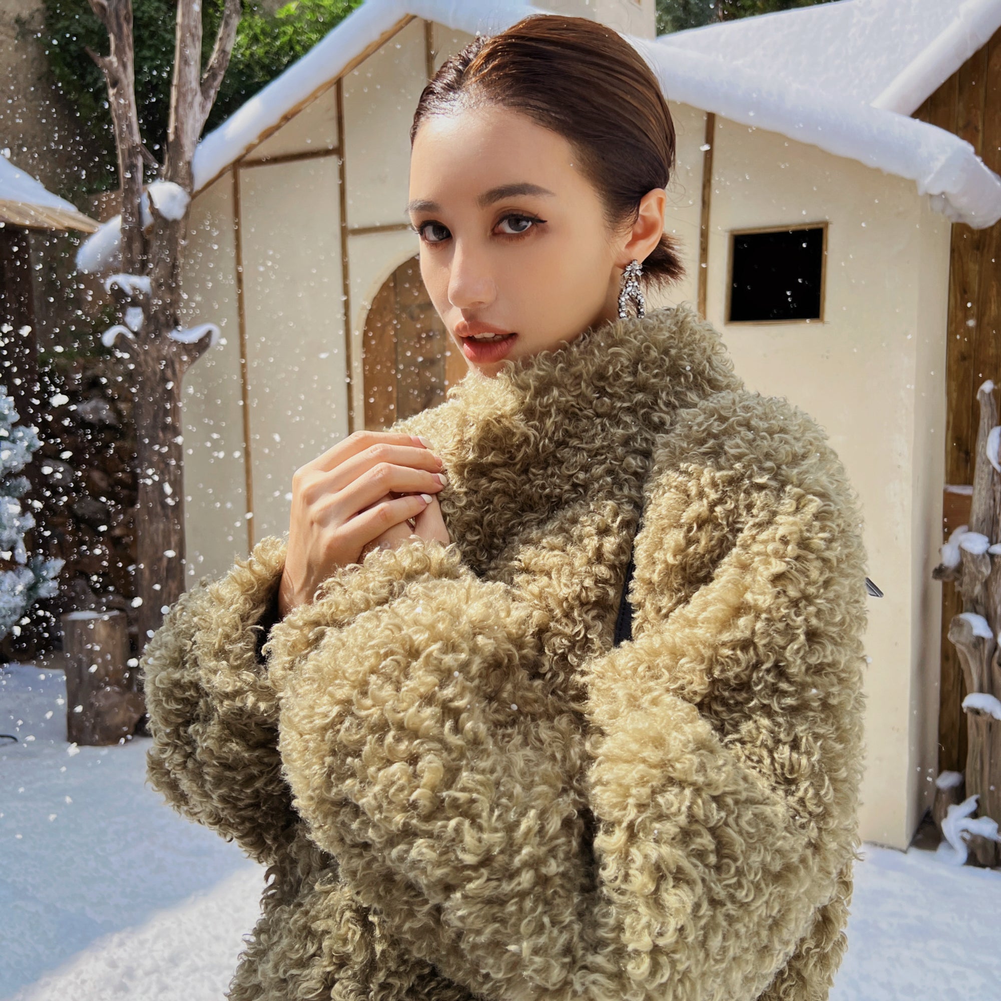 A woman wearing a brown furry coat stands in front of a snowy cabin, with snowflakes falling around her.