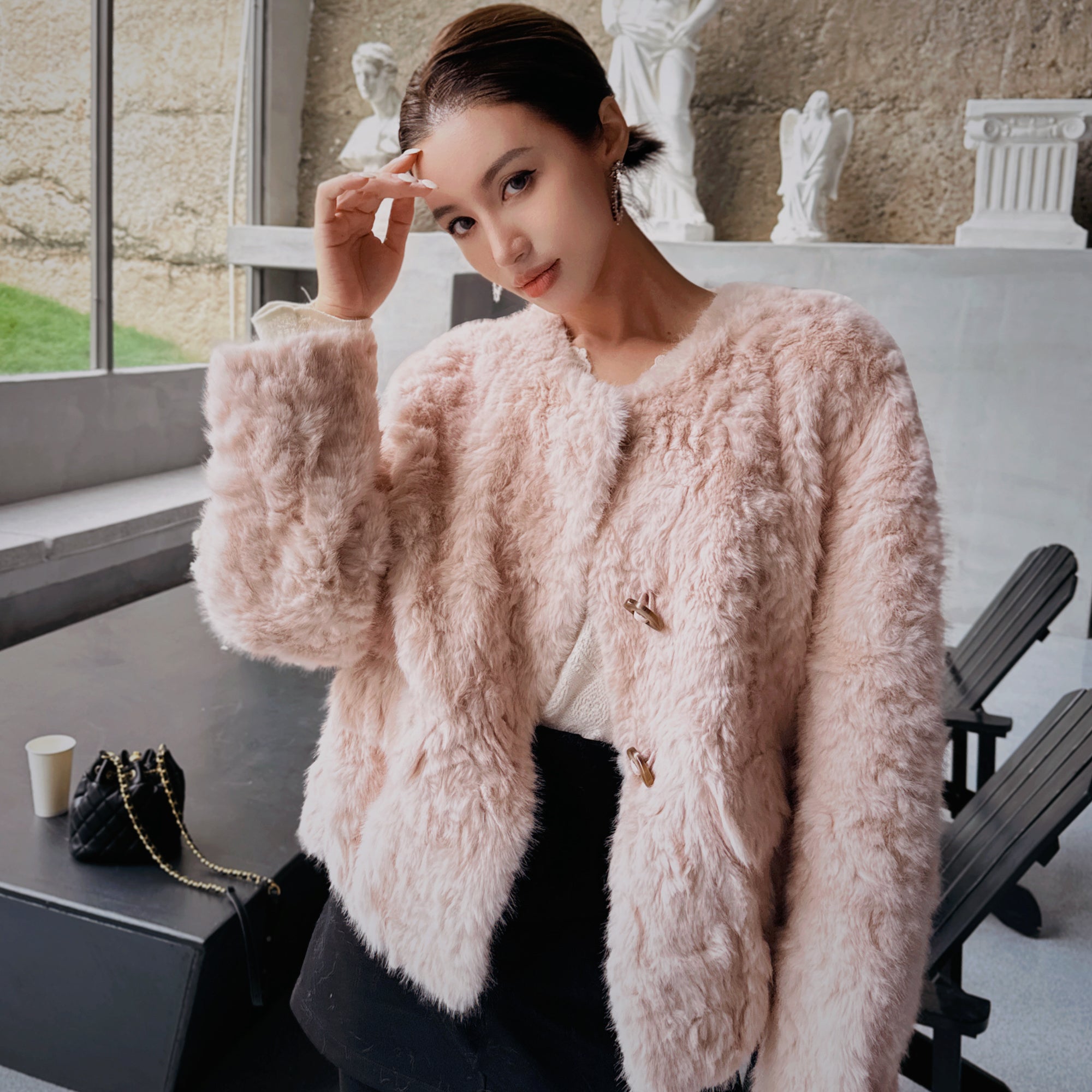 A woman wearing a pink fur coat stands in a room with a window and white statues in the background.