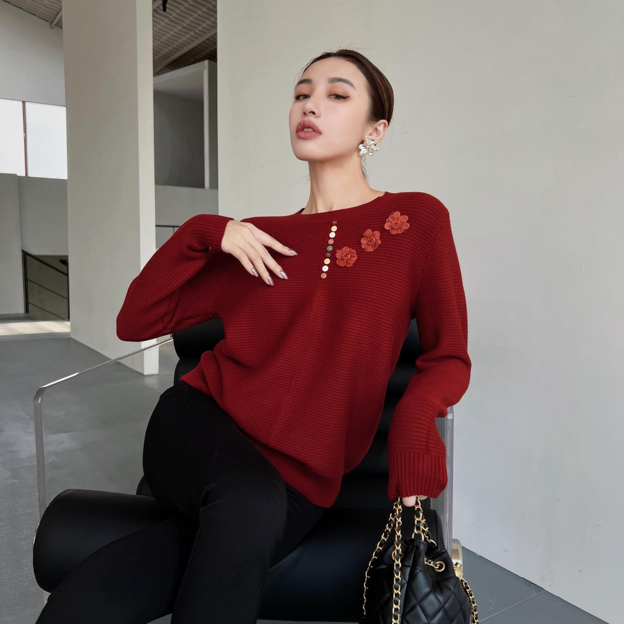 A woman wearing a red sweater with flower designs is sitting on a chair, holding a black purse.