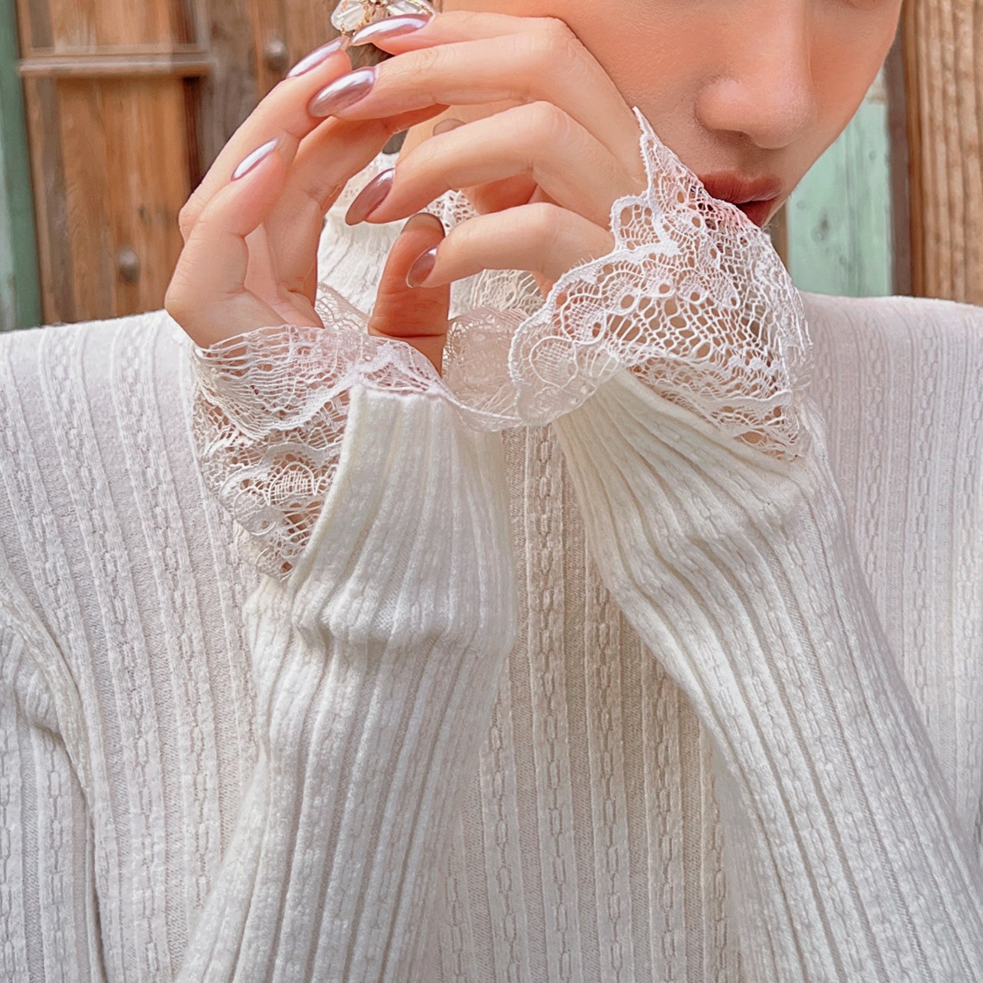 A close-up of a person's hands wearing a white lace-trimmed sweater, with their fingers gently touching their face.