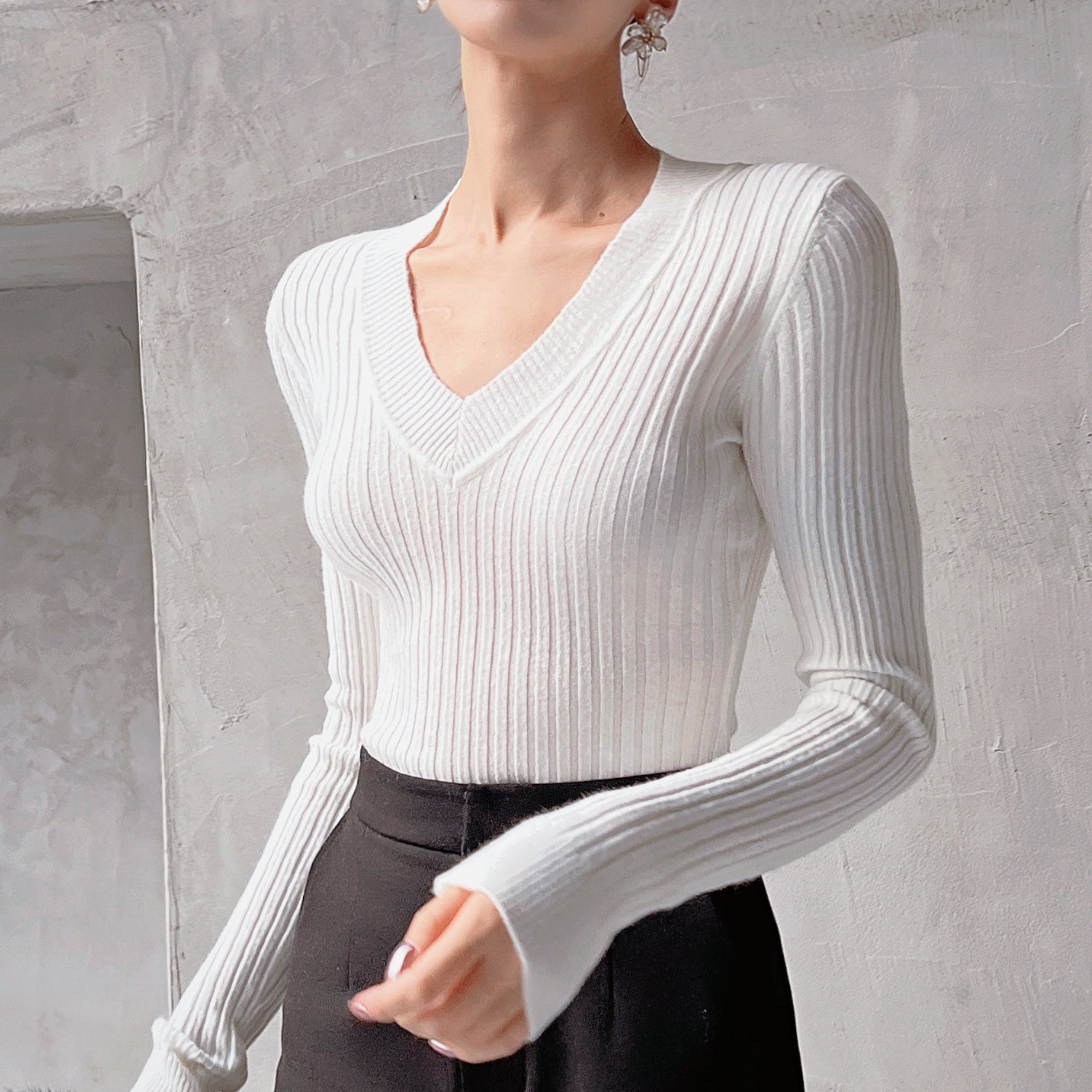 A woman wearing a white v-neck sweater with long sleeves, standing against a plain white wall.