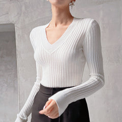 A woman wearing a white v-neck sweater with long sleeves, standing against a plain white wall.