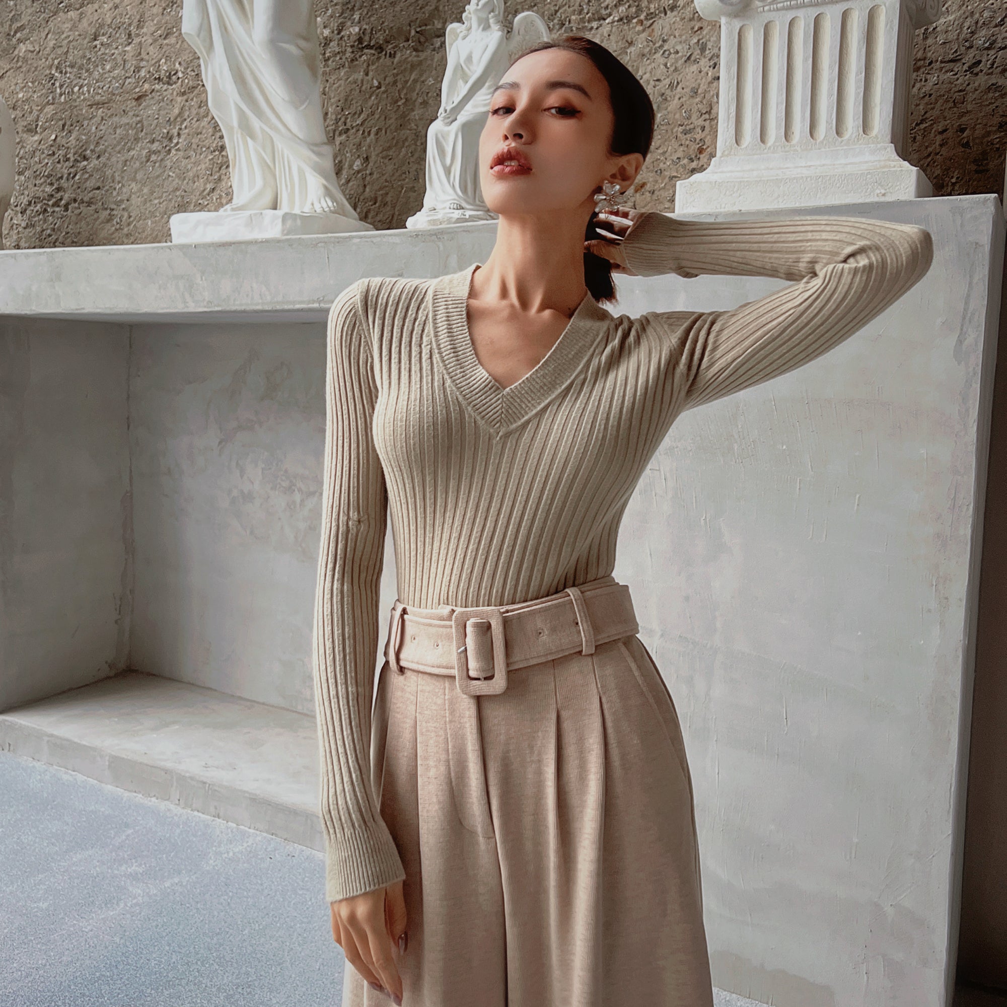 A woman wearing a beige sweater and a beige skirt stands in front of a white marble wall, with a statue visible behind her.