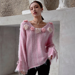 A woman wearing a pink sweater with pink flowers on it, posing in front of a white wall.