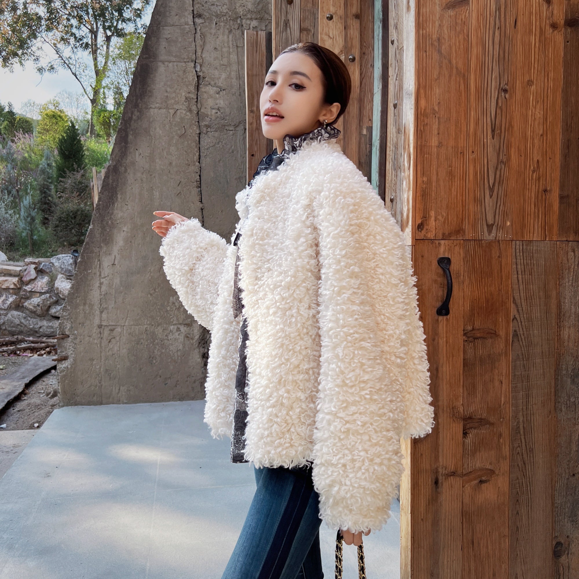 A woman wearing a white fluffy coat stands in front of a wooden structure, with trees visible in the background.
