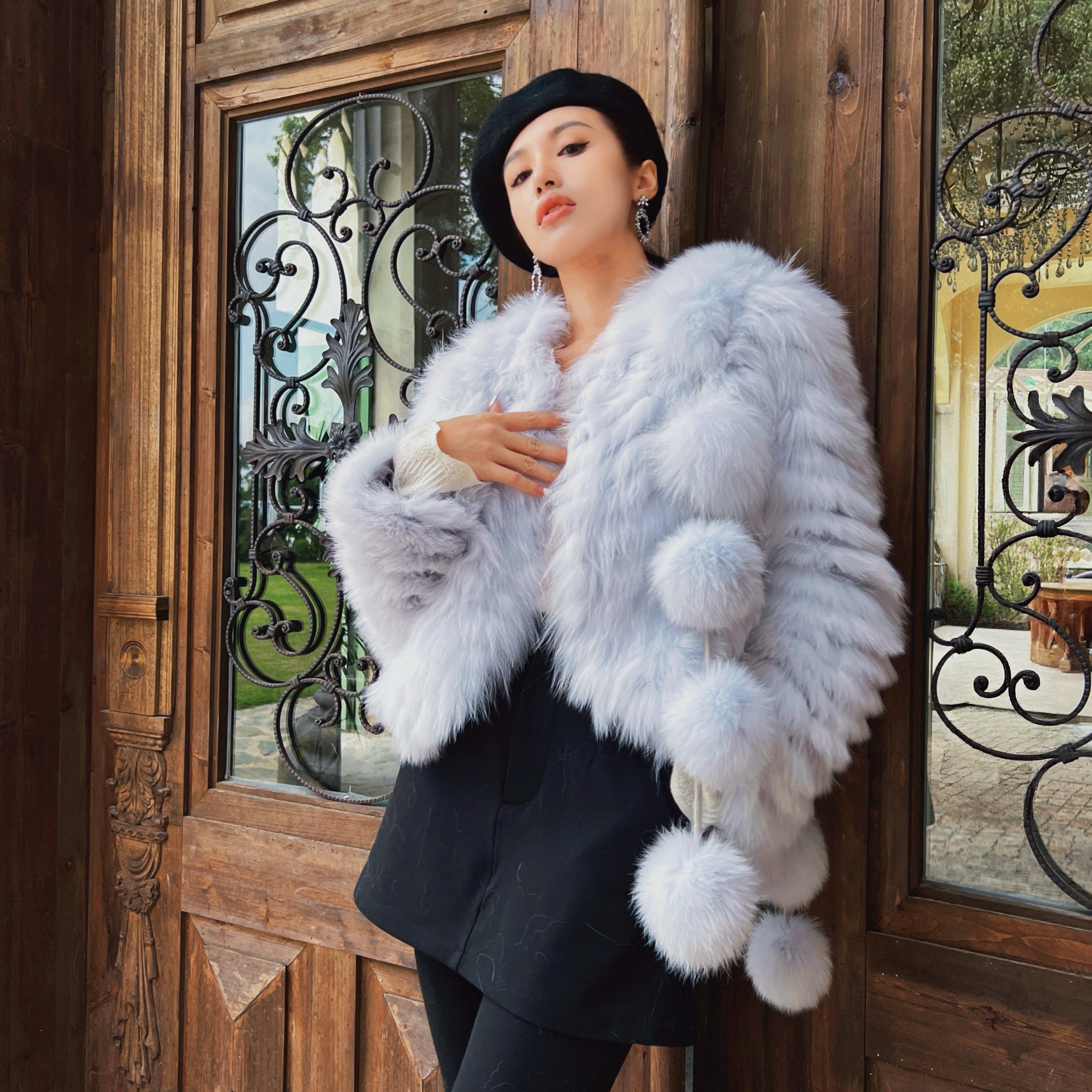 A woman wearing a white fur coat and black skirt stands in front of a wooden door with an ornate wrought iron design.